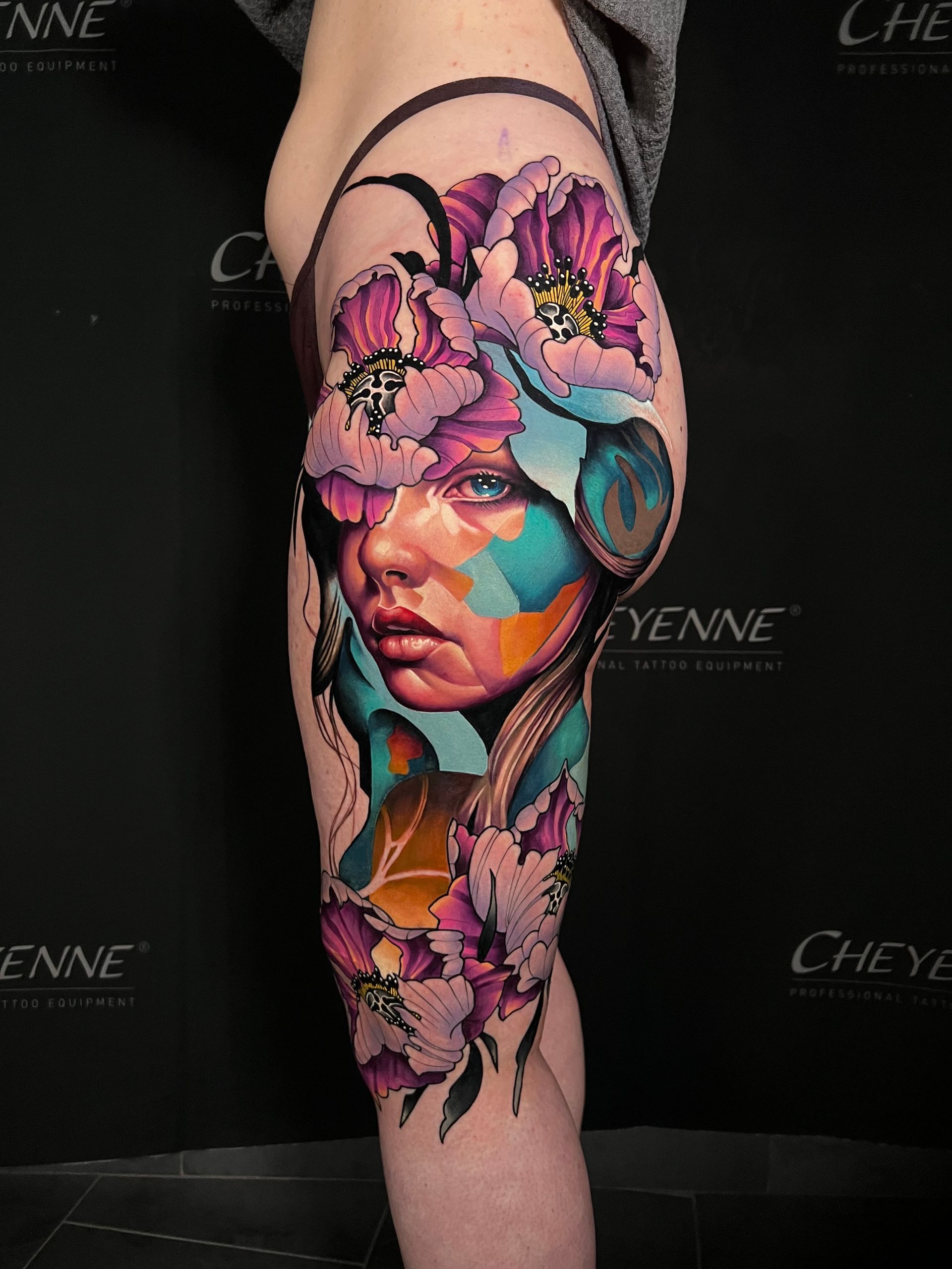 Vibrant portrait tattoo on a person's thigh featuring a woman's face partially obscured by purple and pink flowers, with colorful abstract elements.