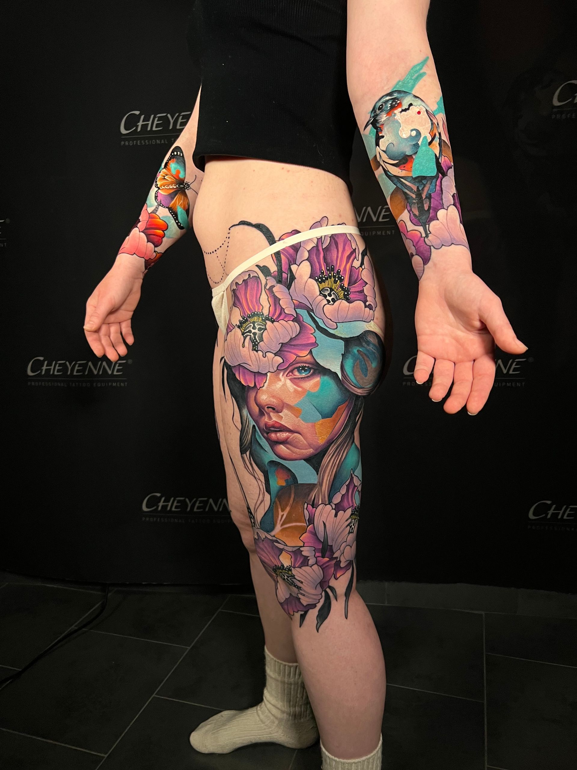 Woman with vibrant floral and portrait tattoos on arms, hip, and leg, against a dark background.
