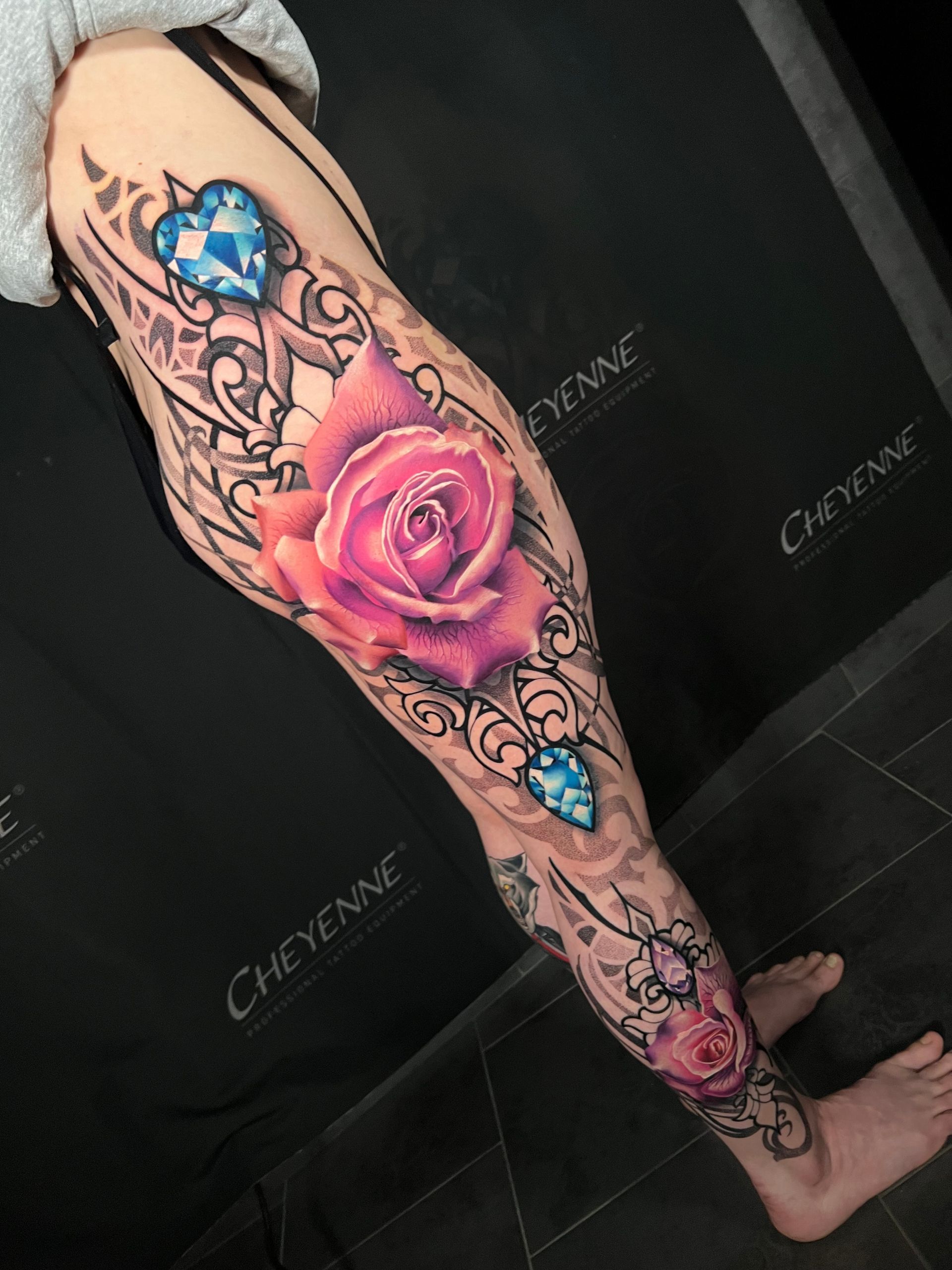 Leg tattoo featuring a large pink rose and blue gemstones framed by ornate black line art. The tattoo extends from the upper thigh to the foot.