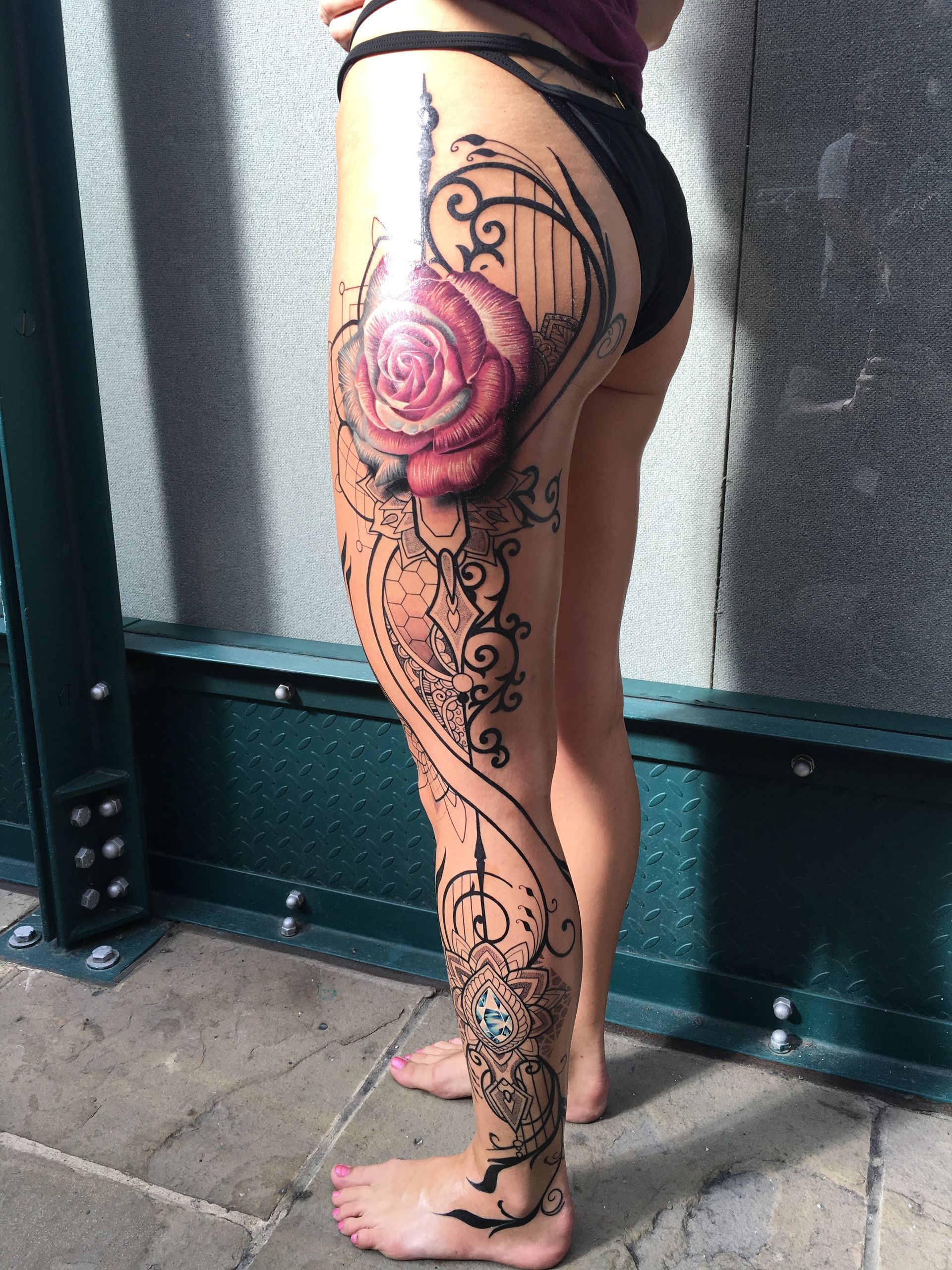 Woman's leg with large rose tattoo, surrounded by black ornate filigree, standing outdoors.