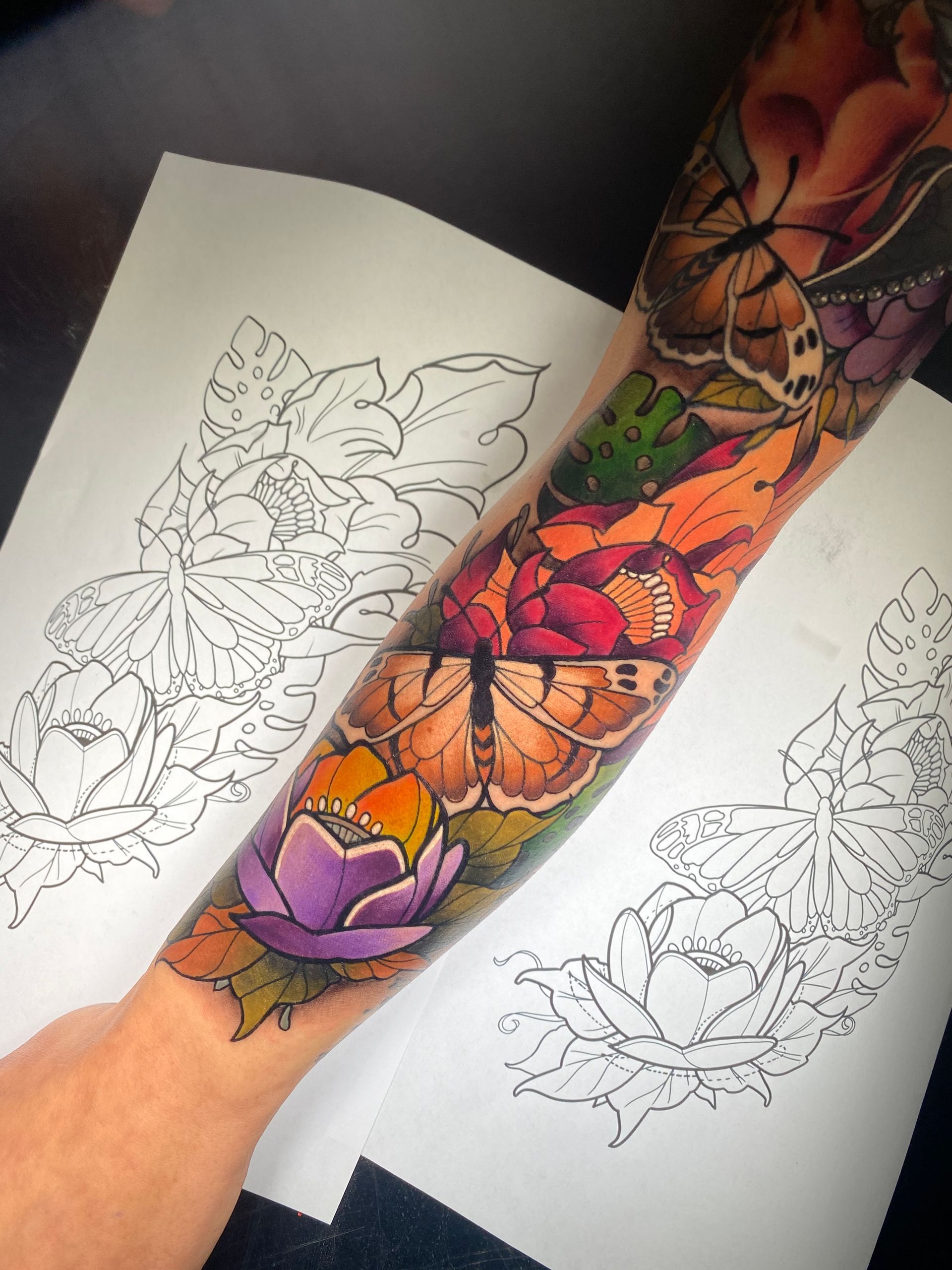 Colorful floral and butterfly tattoo on a forearm, next to outline sketches on paper.