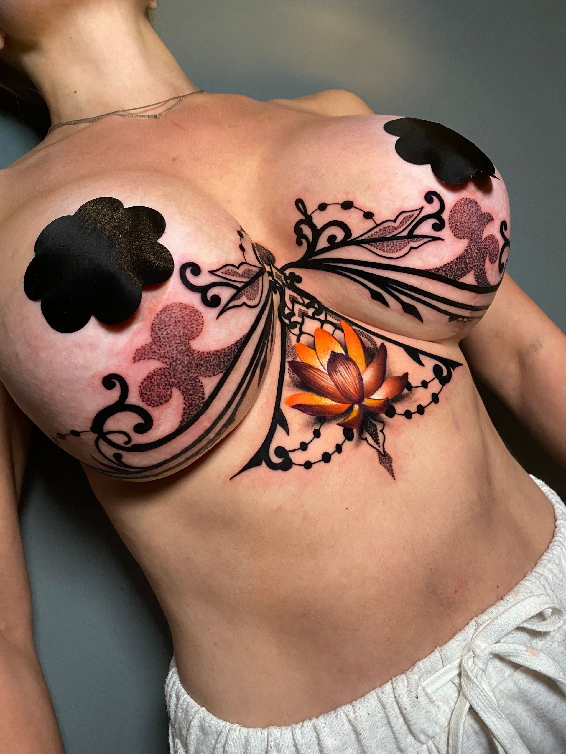 Woman with a chest tattoo of a lotus flower and decorative black and pink embellishments. Nipple covers are worn.