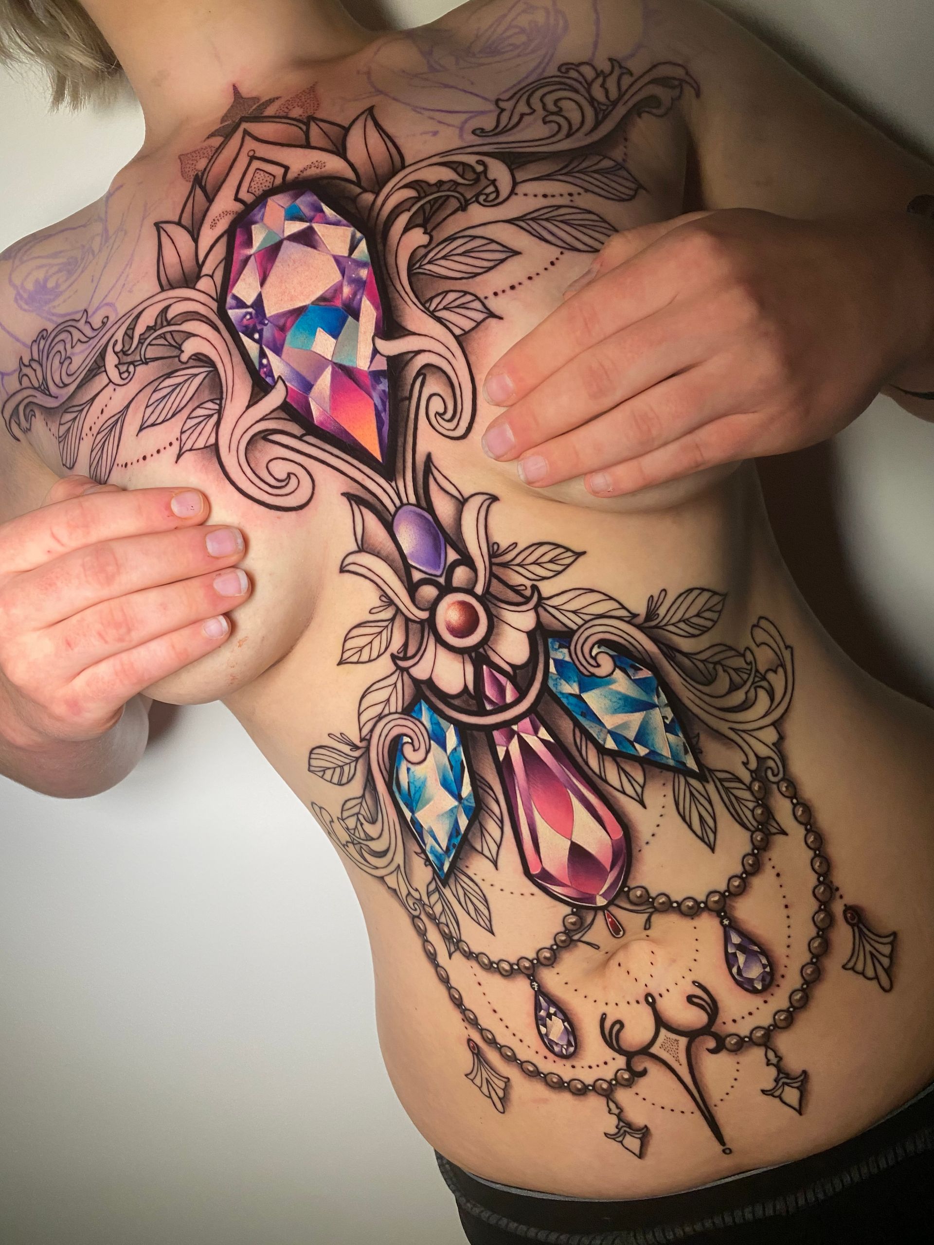 Woman with chest tattoo of a jeweled pendant, hands covering breasts. Tattoo features colorful gems, black line work, and elaborate detailing.