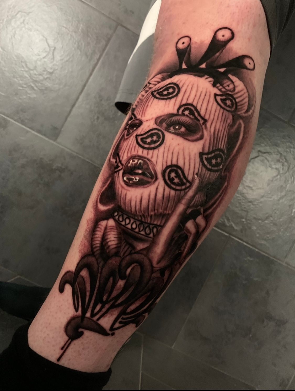 Tattoo on a leg: a person wearing a bandana, with exposed eyes and mouth, holding a phone; dark shading.