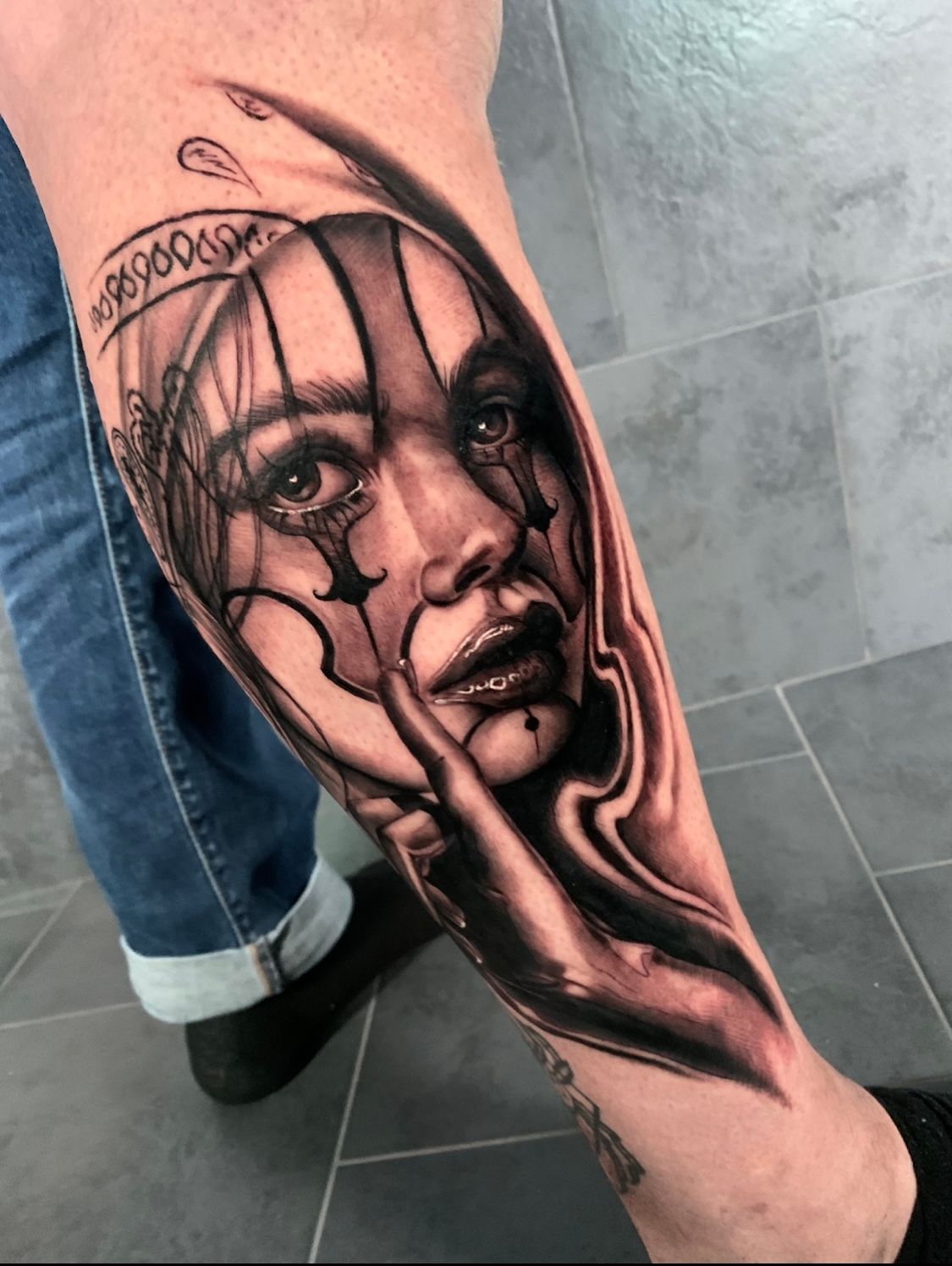 Black and white tattoo of a woman with clown makeup on a leg. She is holding her finger to her lips with a tear falling from her eye.