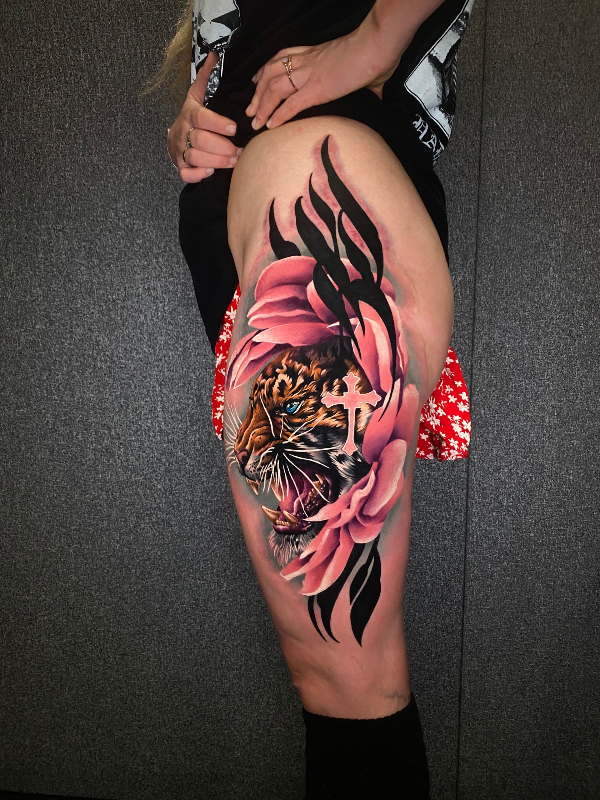 A colorful tattoo of a tiger, pink flower, and cross on a person's upper thigh. Black accents and shadows.