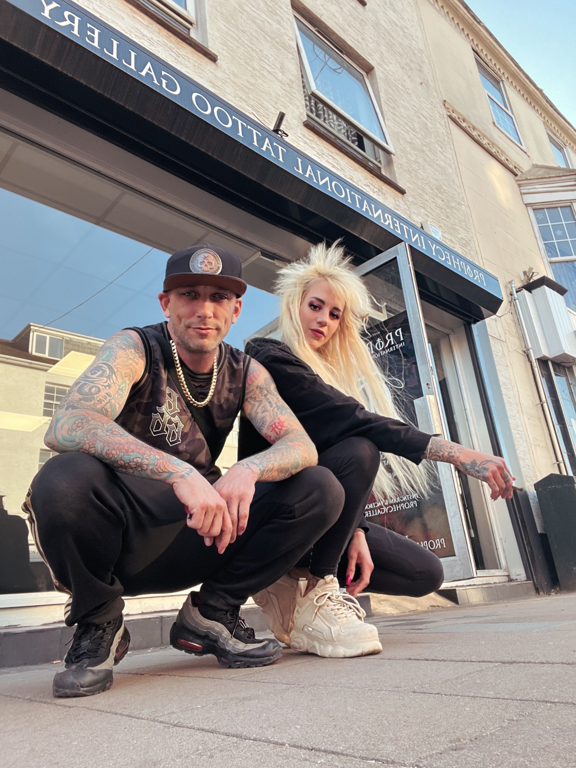 Man and woman with tattoos pose crouching outside a tattoo parlor. The man wears a cap and gold chain. The woman has blonde hair and a black jacket.