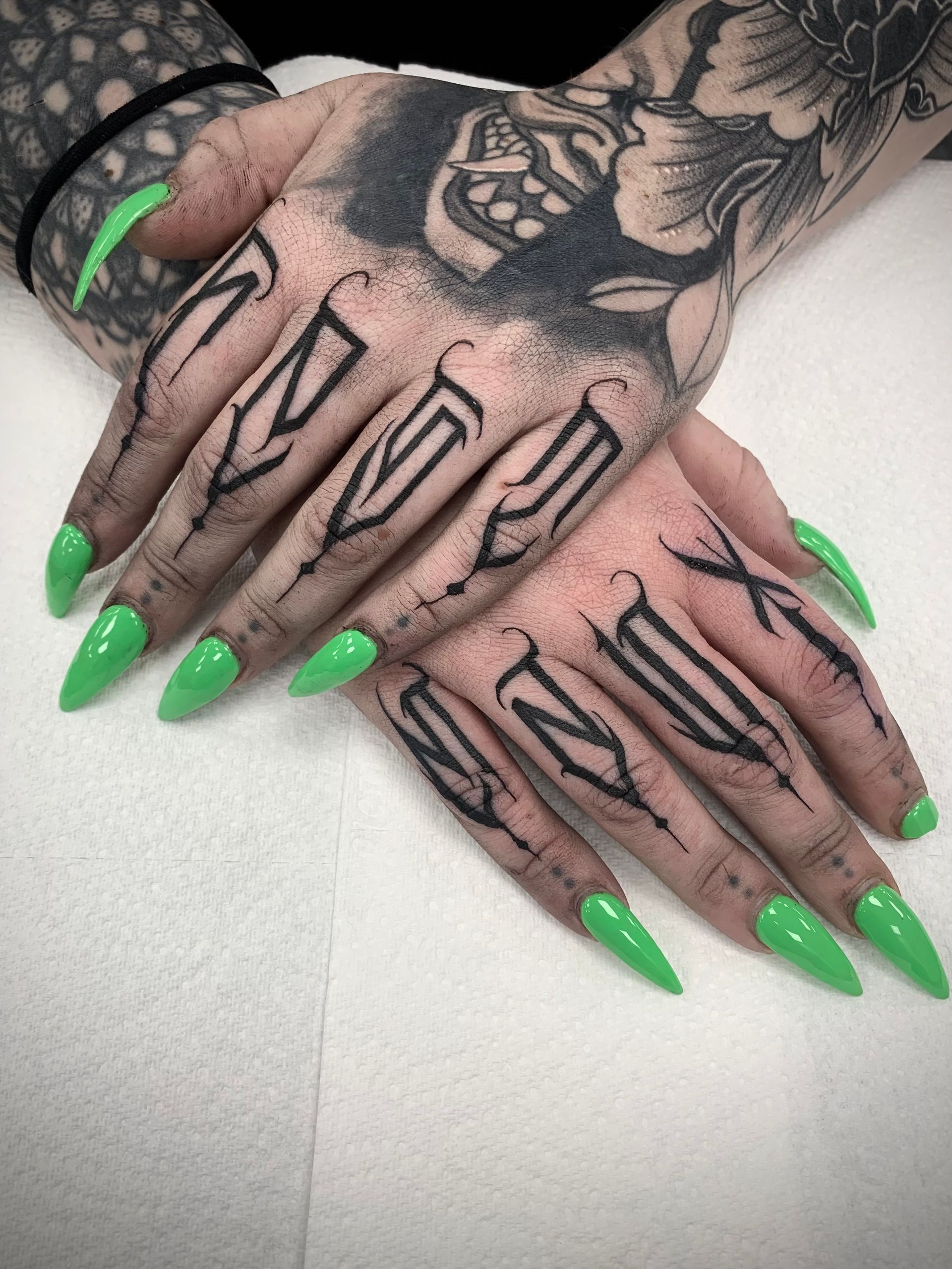 Hands with green stiletto nails, finger tattoos of black stylized letters, and intricate black sleeve tattoo on the left arm.