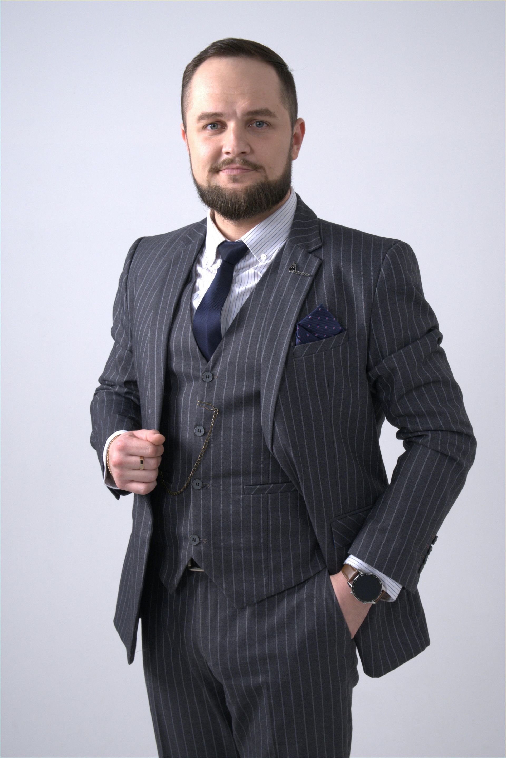 A person in a charcoal pinstriped three-piece suit and tie stands against a plain light gray background.