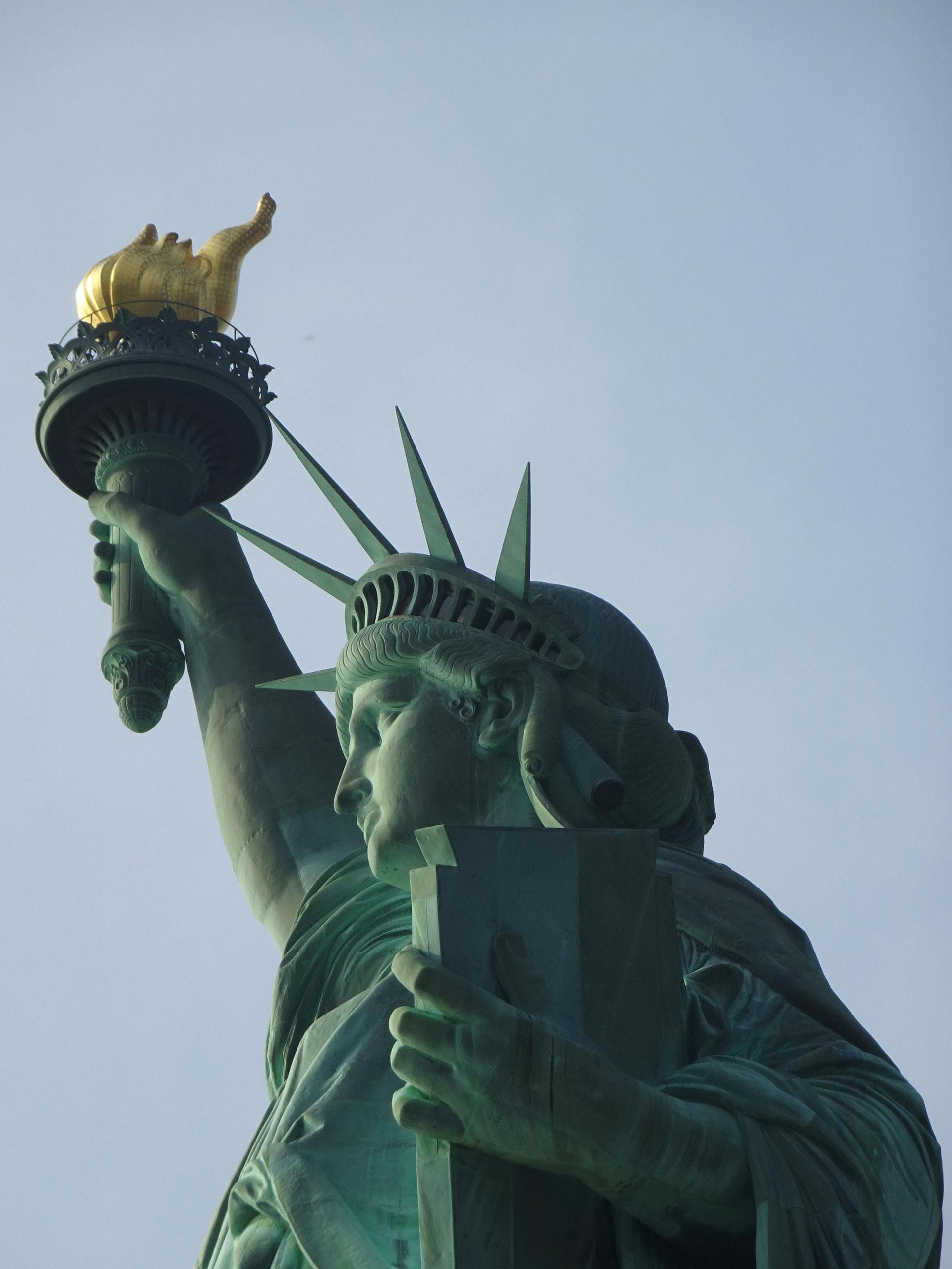 A low-angle view of the Statue of Liberty holding her torch against a pale blue sky.