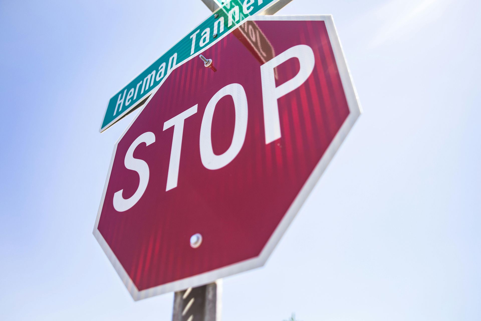 A red octagonal stop sign with white text, mounted on a pole below a street sign labeled Herman Tanner.