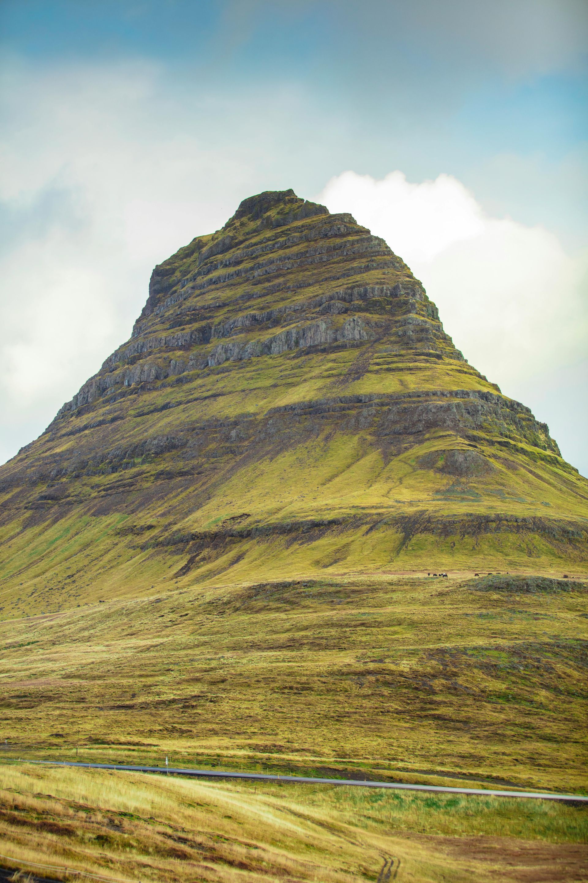 Kirkjufell mountain in Iceland, featuring distinct horizontal rock layers covered in green grass under a cloudy sky.