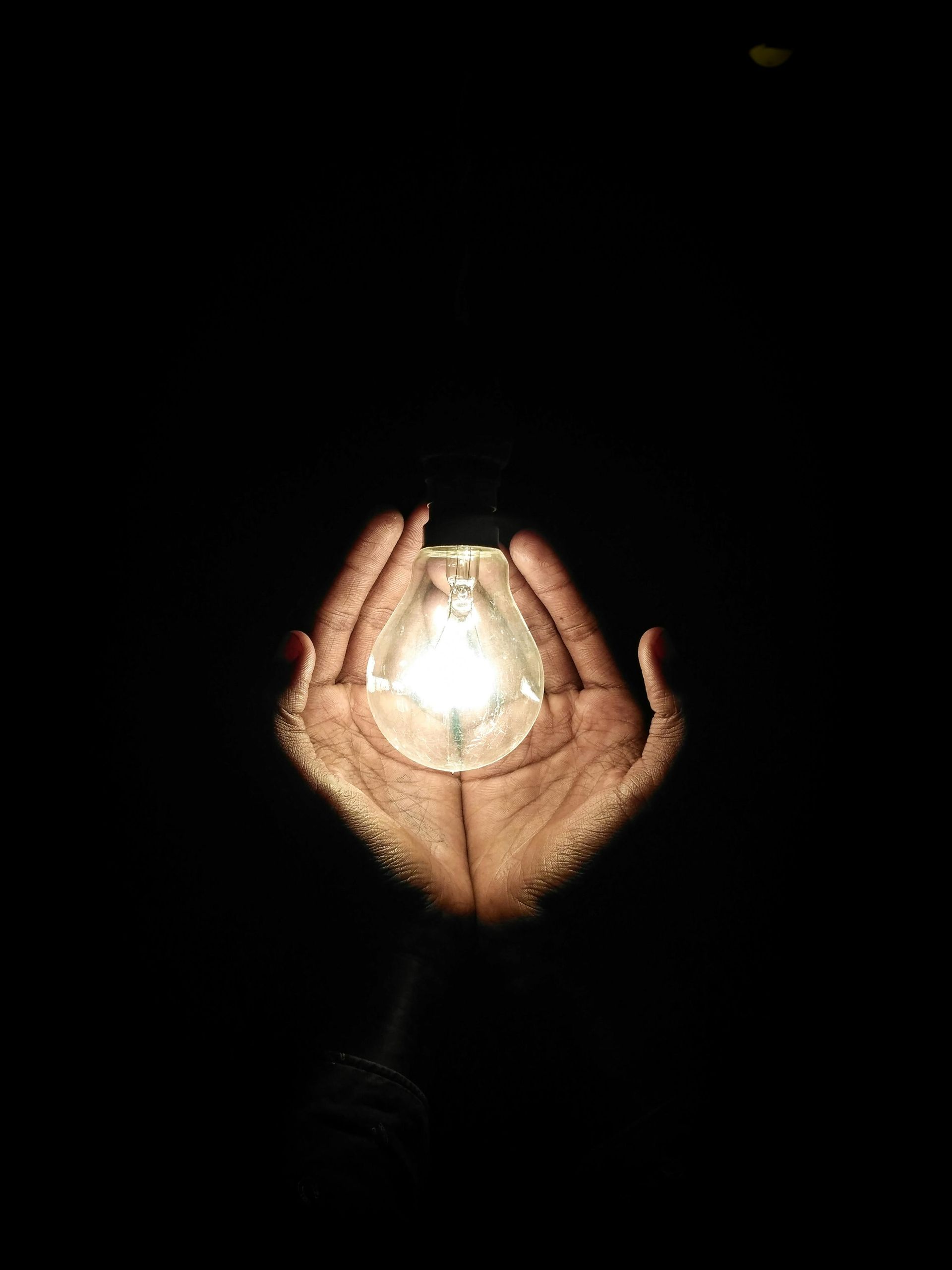 Hands cupped around a glowing light bulb against a solid black background.