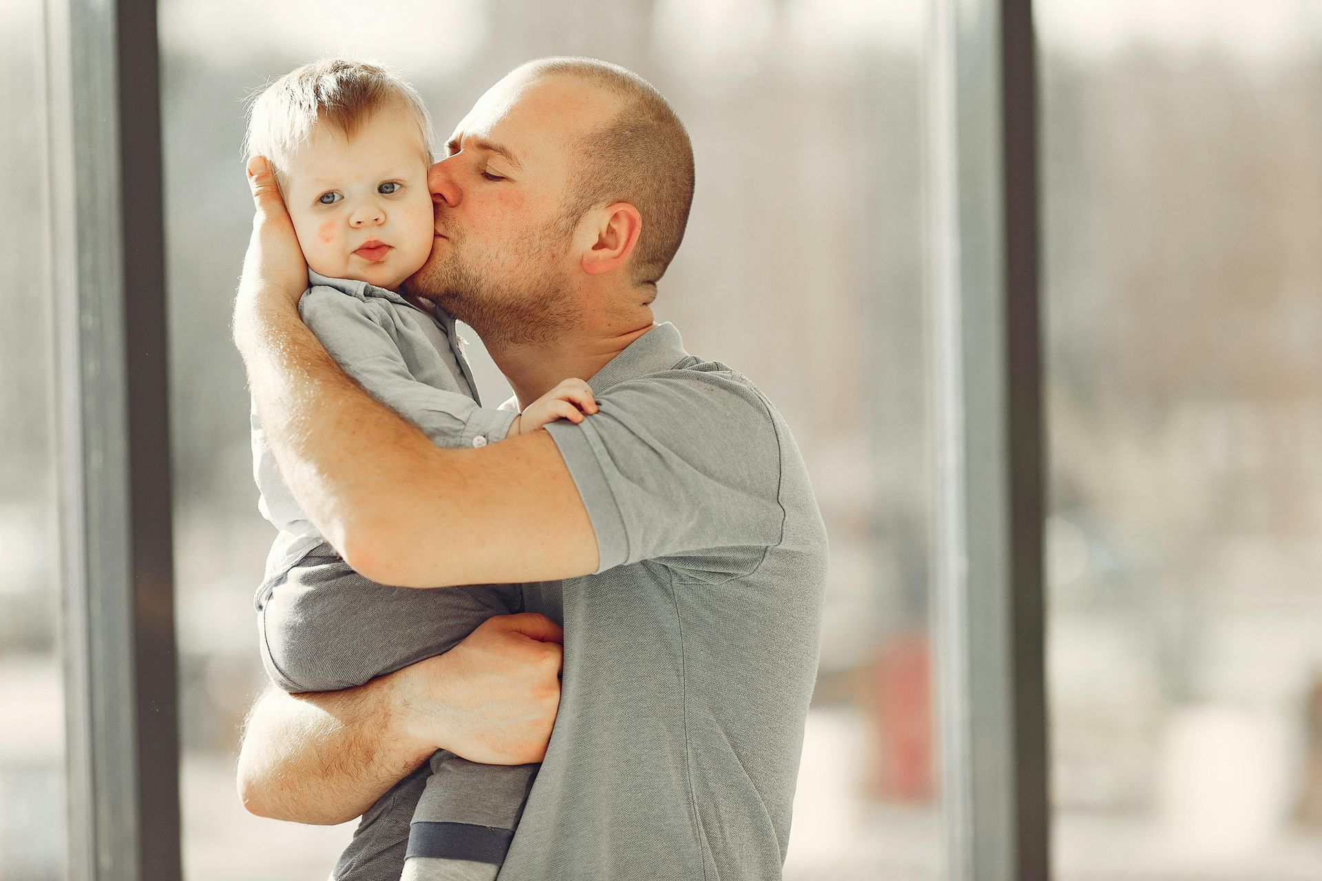 A man in a grey shirt holding a small child and kissing their cheek near a large window.