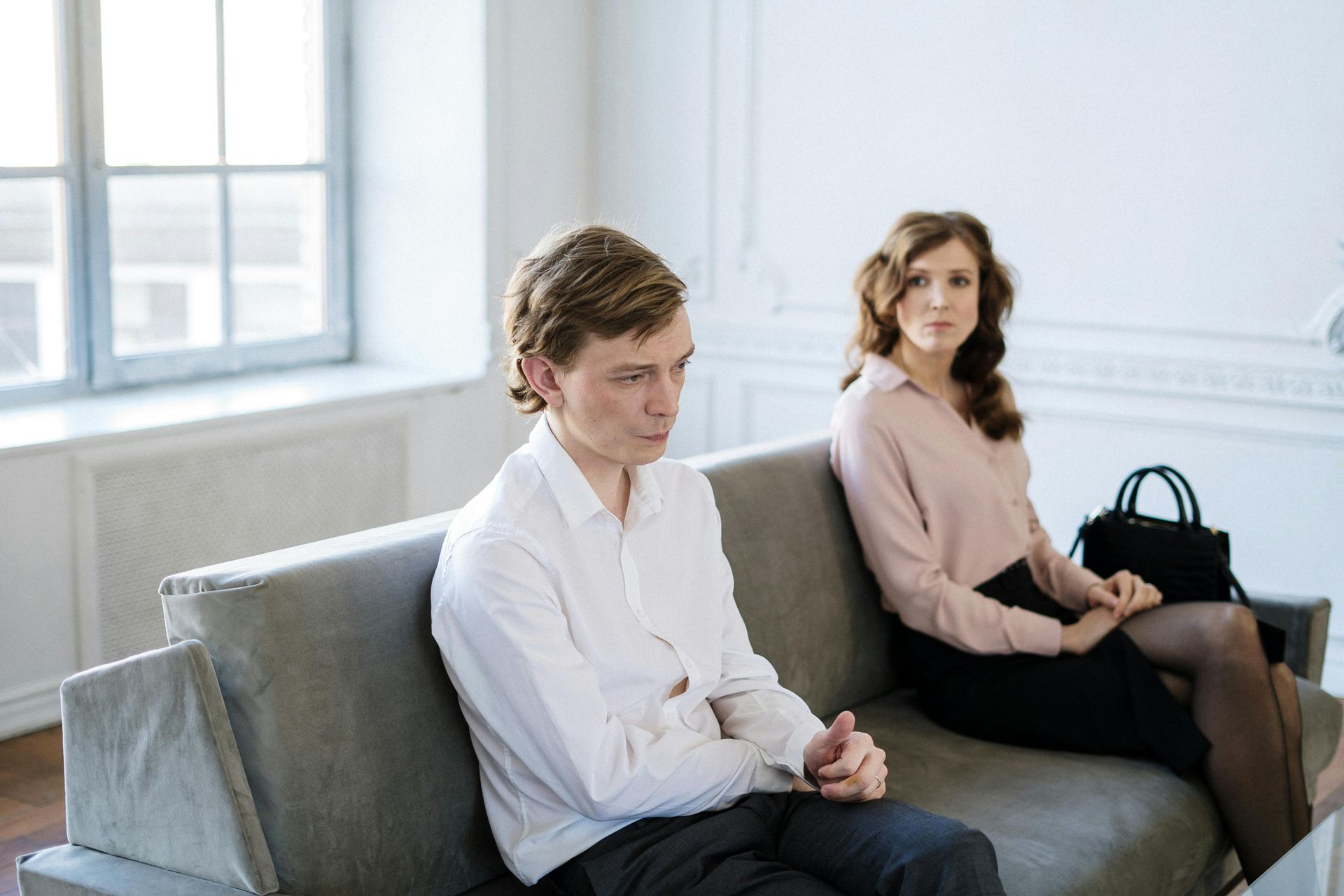 Two people sit on a grey sofa in a brightly lit, minimalist room. They look away from each other with neutral expressions.