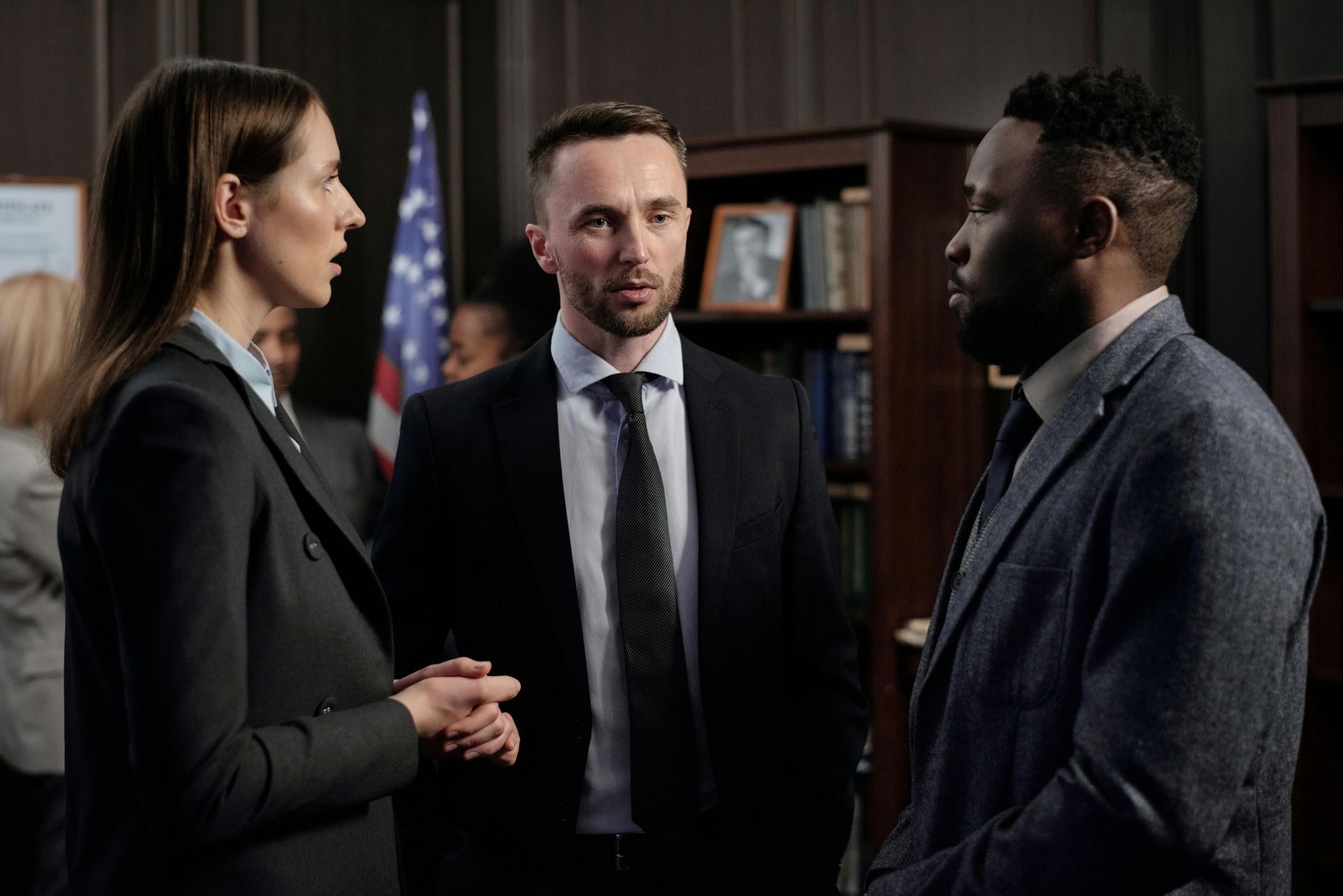 Three professionals in formal business attire talk while standing in an office with a bookshelf and a flag in the background.
