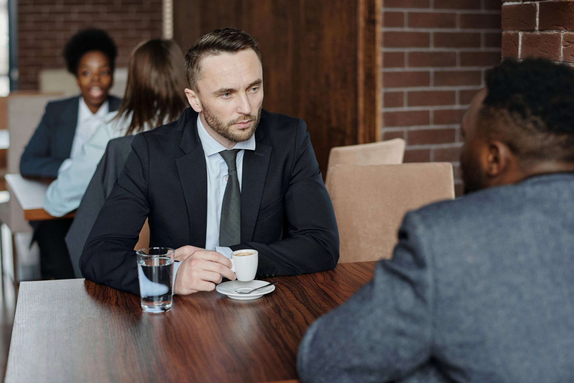 Two professionals in business attire hold a meeting over coffee at a wooden table in a cafe with other people in the back.