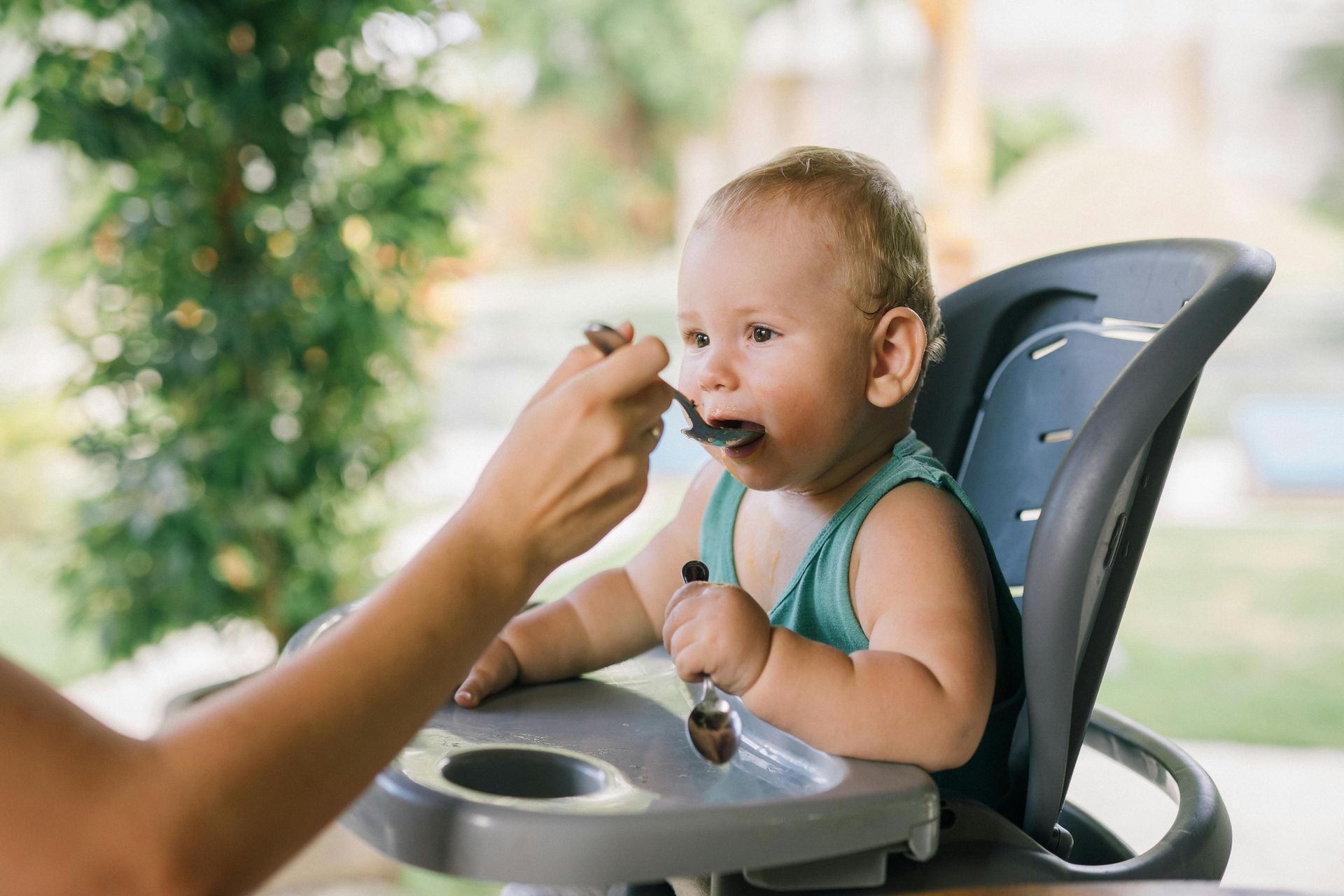 An adult feeds a baby in a high chair using a spoon while the baby holds a second spoon.