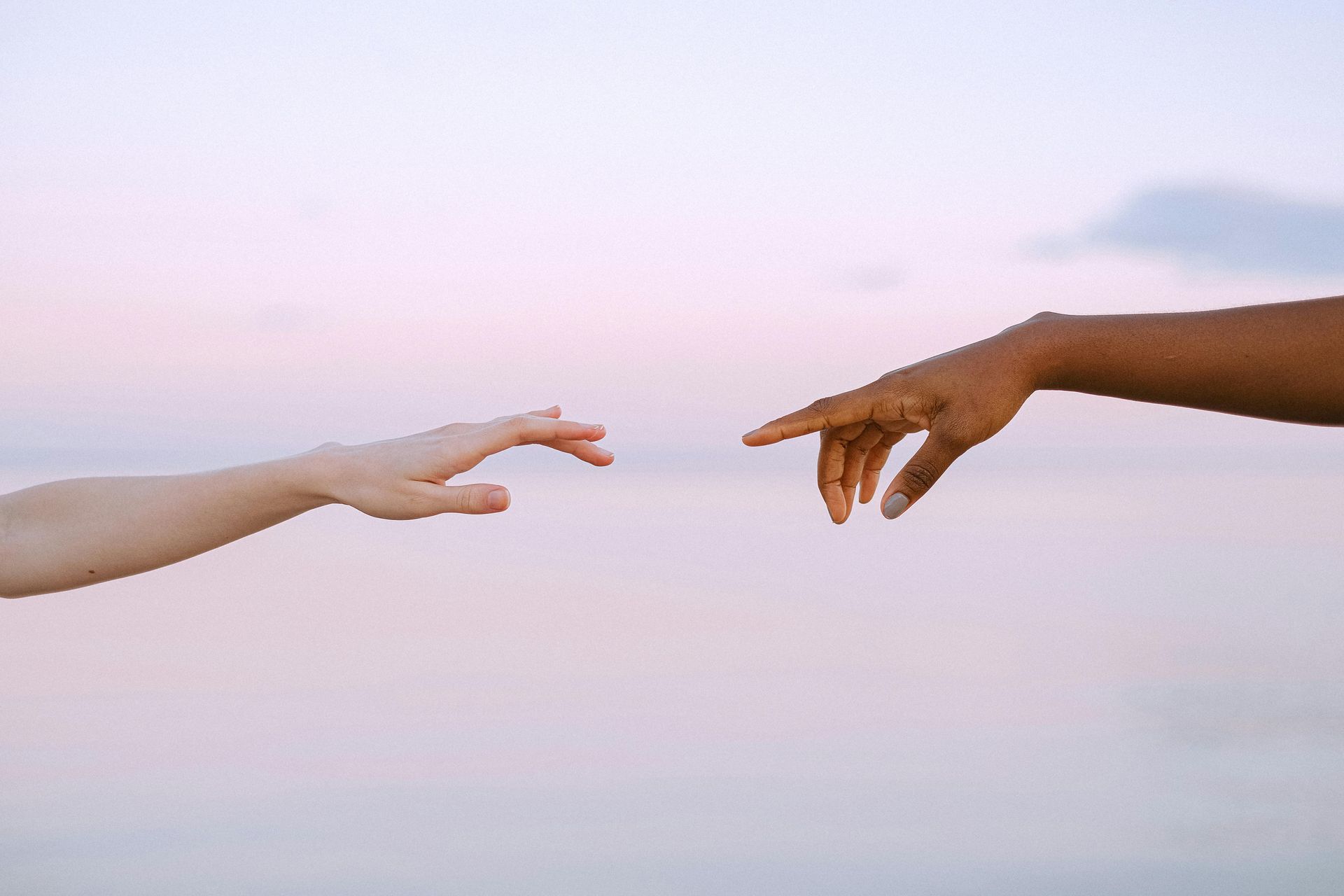 Two hands reaching toward each other against a soft, pastel-colored sky.
