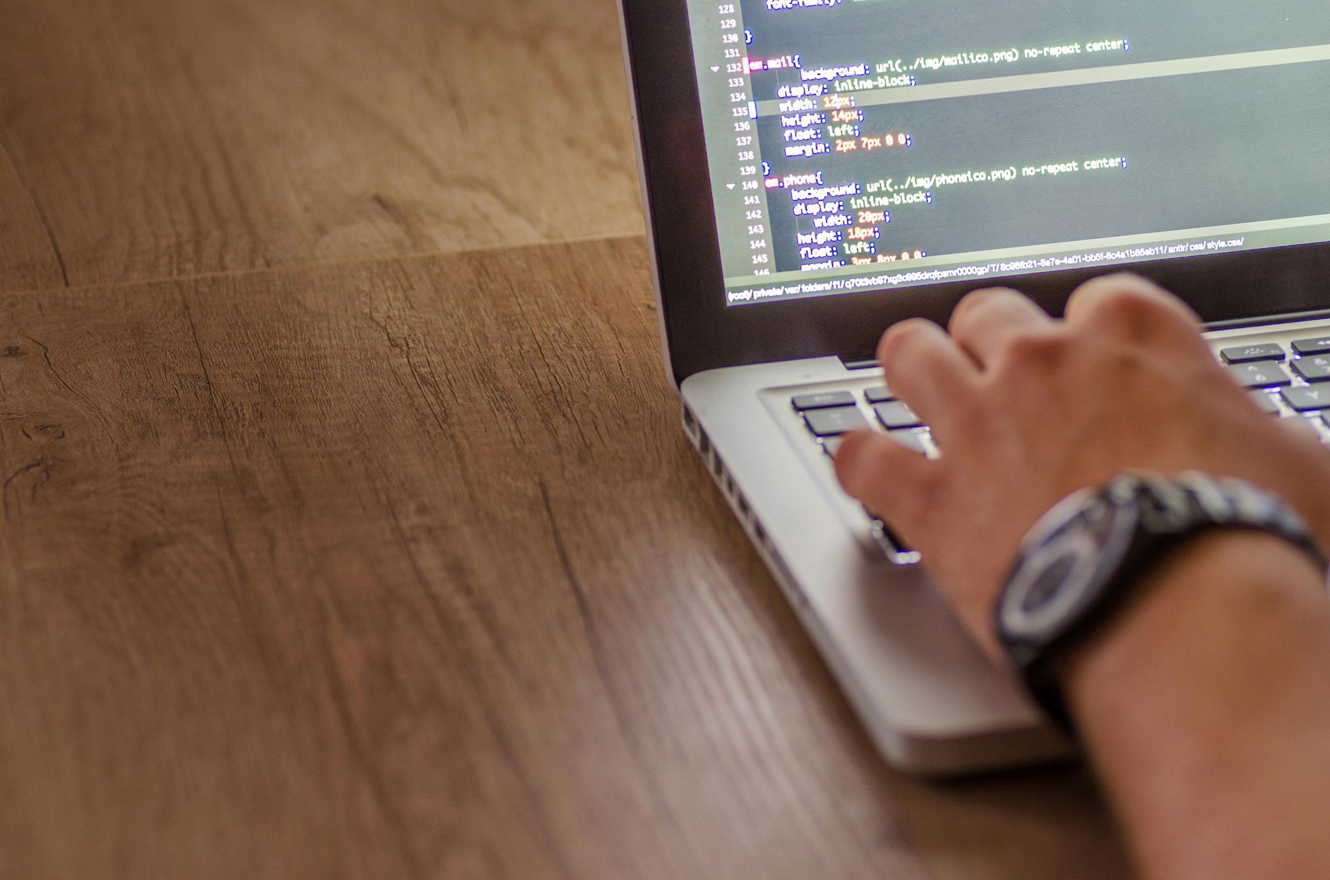 A person with a watch typing on a laptop displaying code, viewed from above on a wooden desk.