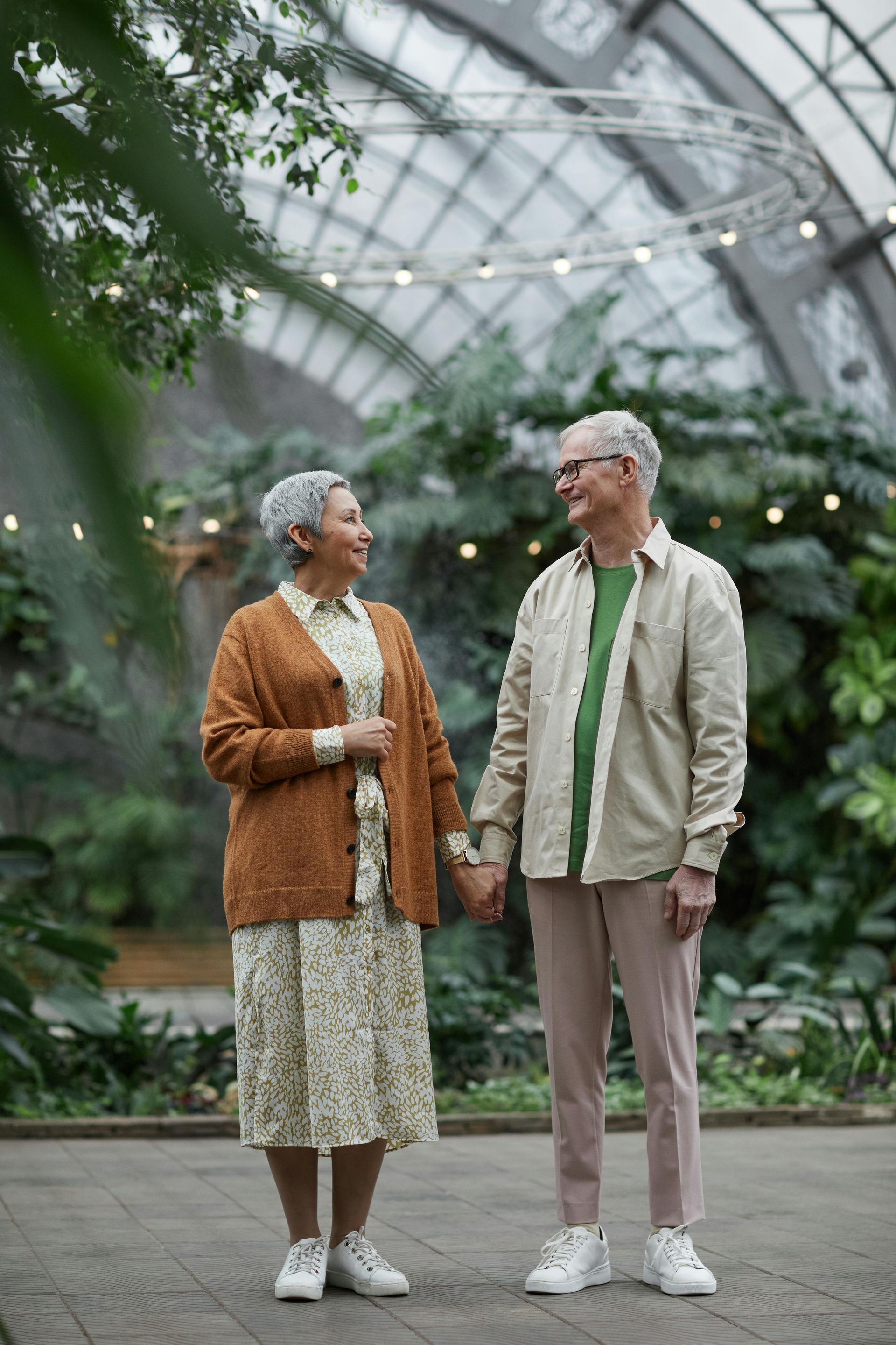 Two people holding hands, standing together in a lush greenhouse, looking at each other and smiling.