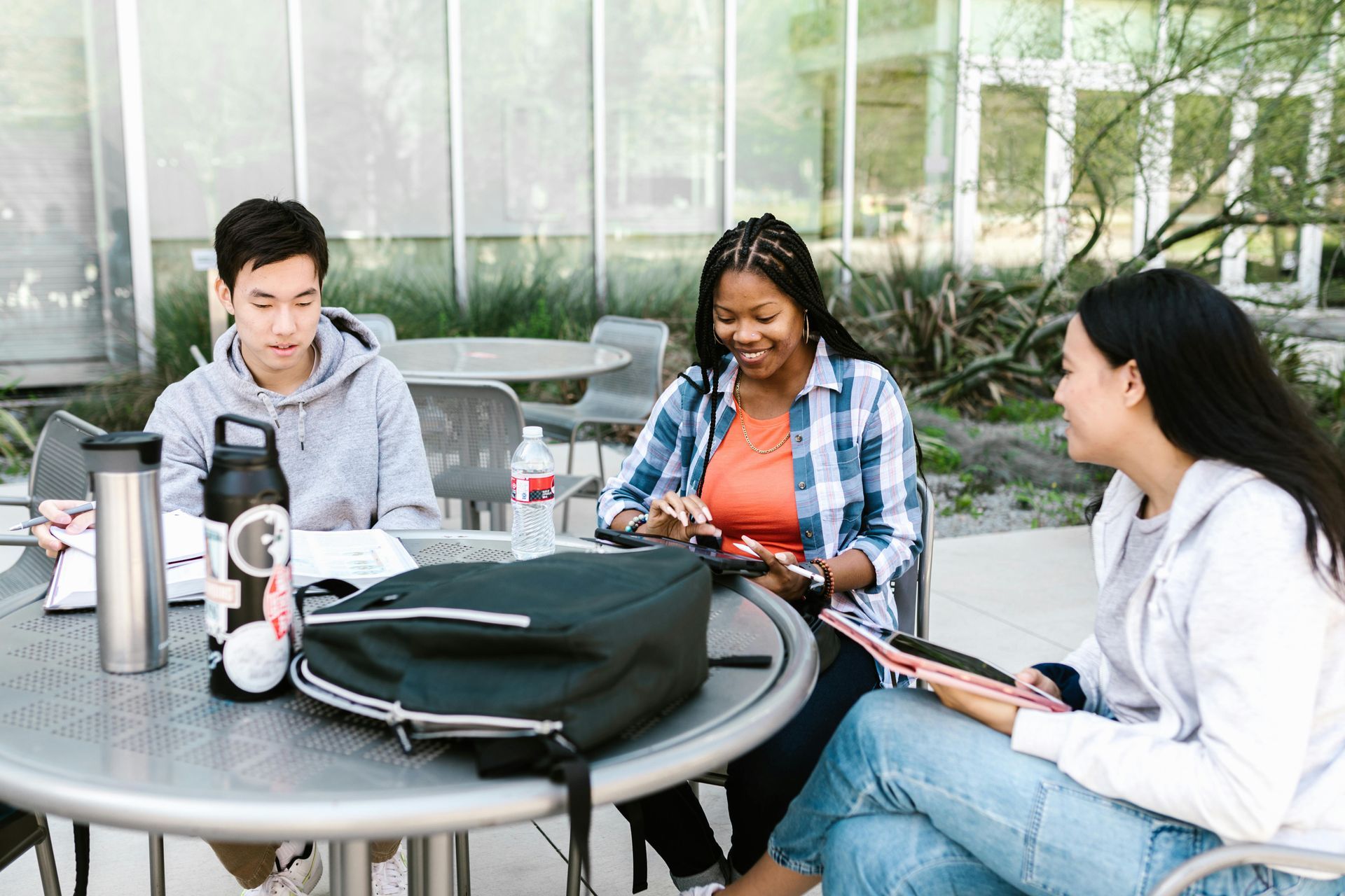 Three students sit around an outdoor metal table studying together, with a black backpack, notebooks, and water bottles.