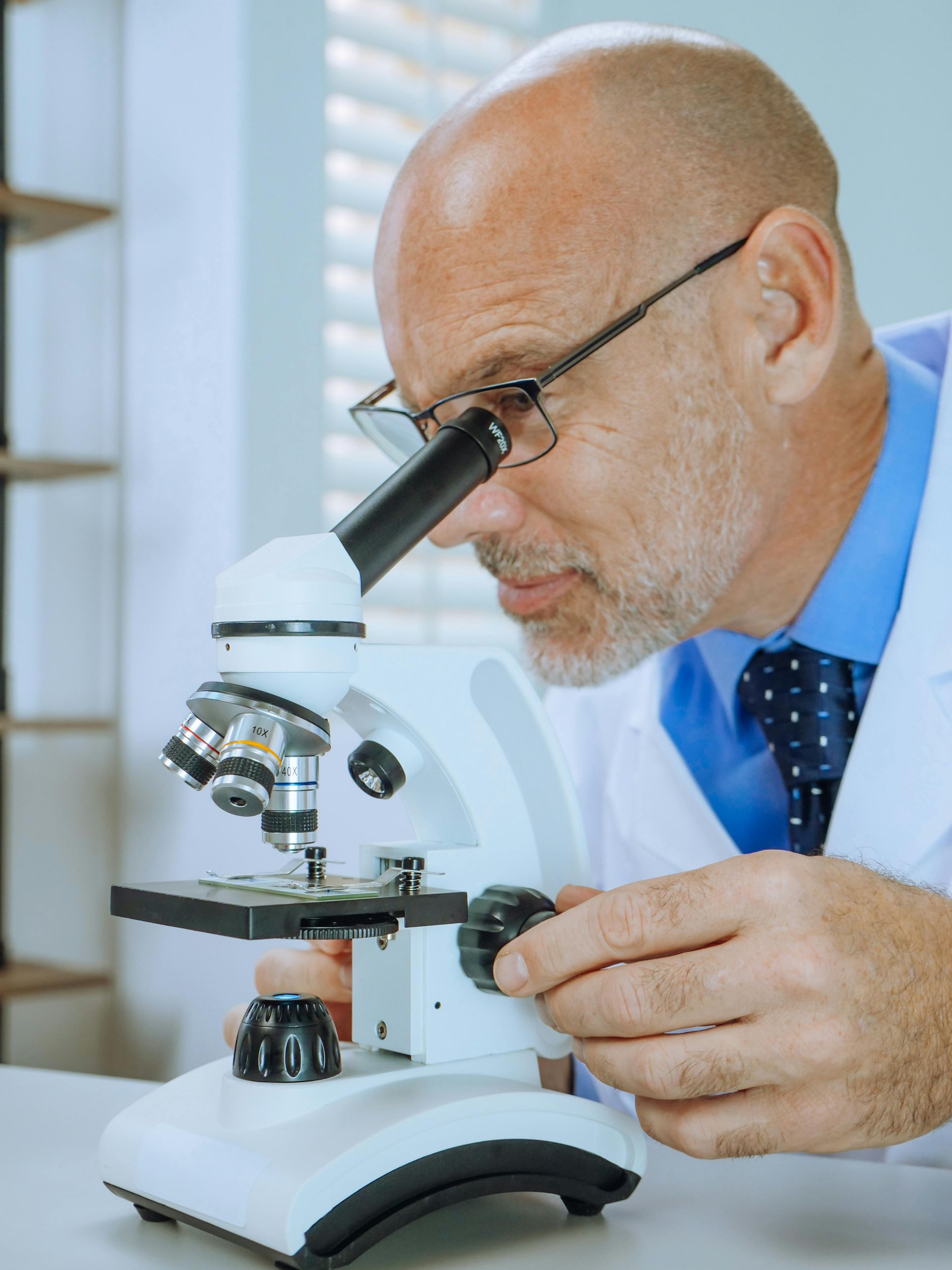 A person wearing a lab coat and glasses adjusts the focus knob on a white microscope.