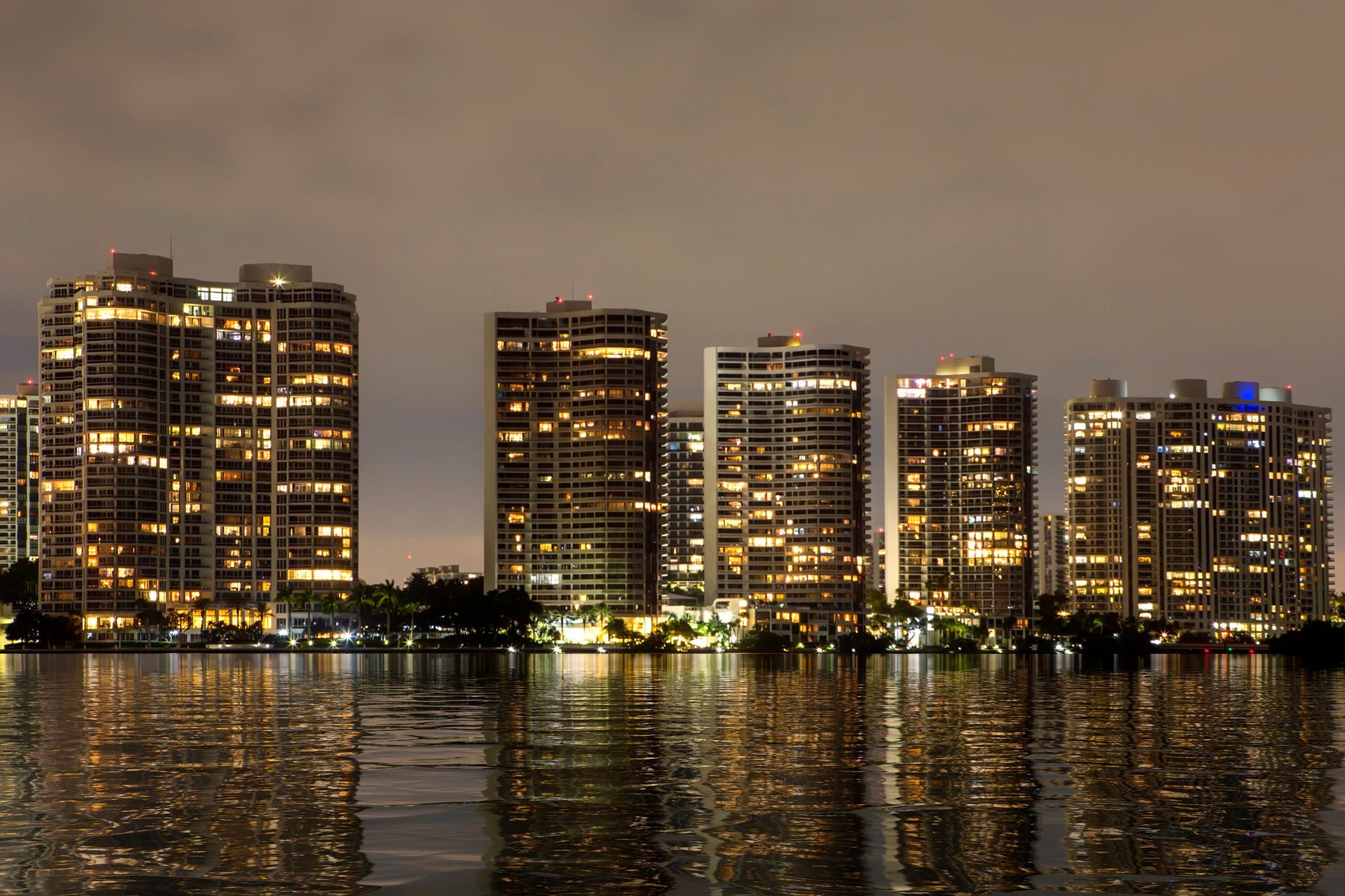 A row of illuminated high-rise apartment buildings reflecting on a dark body of water at night.