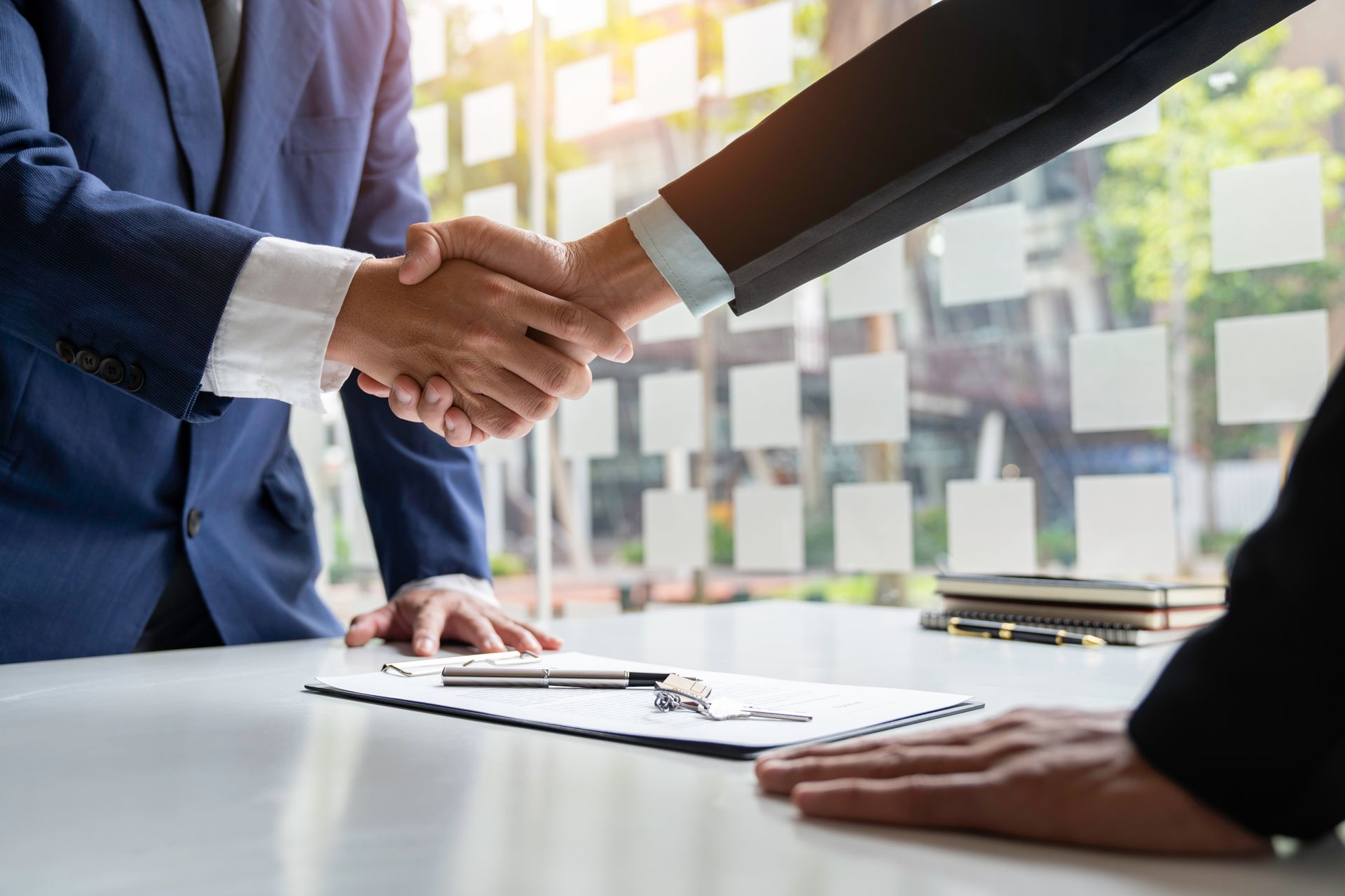 Two business professionals in suits shaking hands over a contract on a desk in a bright office.