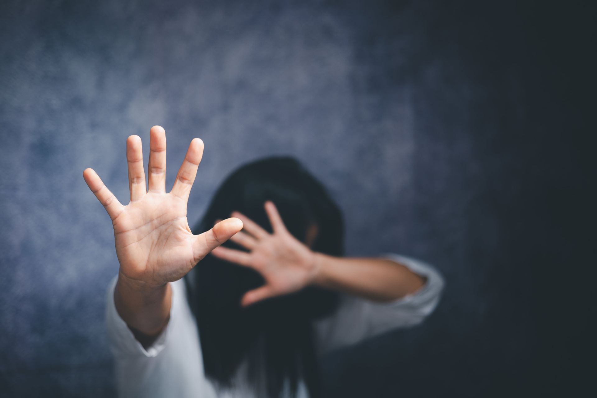 A person in a white top holds their hands up in a defensive or shielding gesture against a textured dark background.