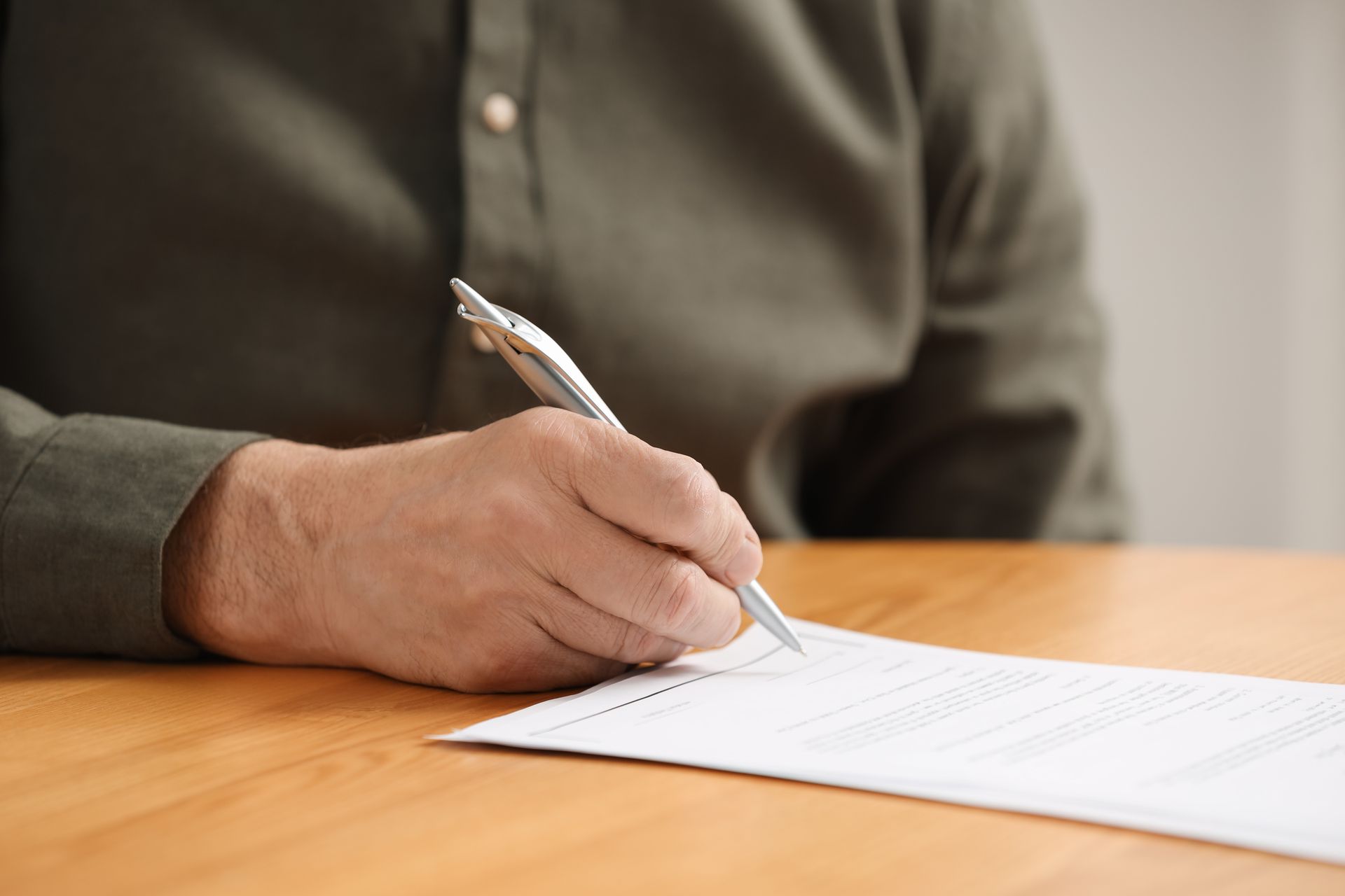 A hand in a long-sleeved green shirt writes on a piece of paper with a silver pen at a wooden table.