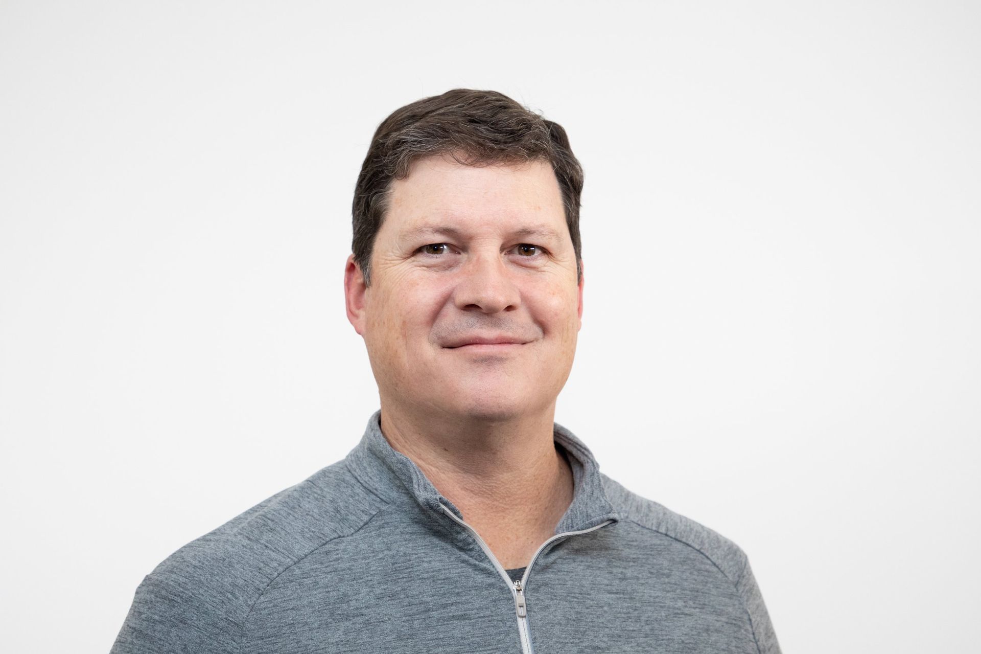 Man in gray zip-up sweater smiles at the camera against a white background.