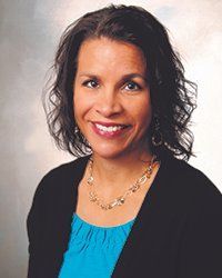 Woman with dark hair smiles, wearing a black sweater over a blue top, and gold necklace.