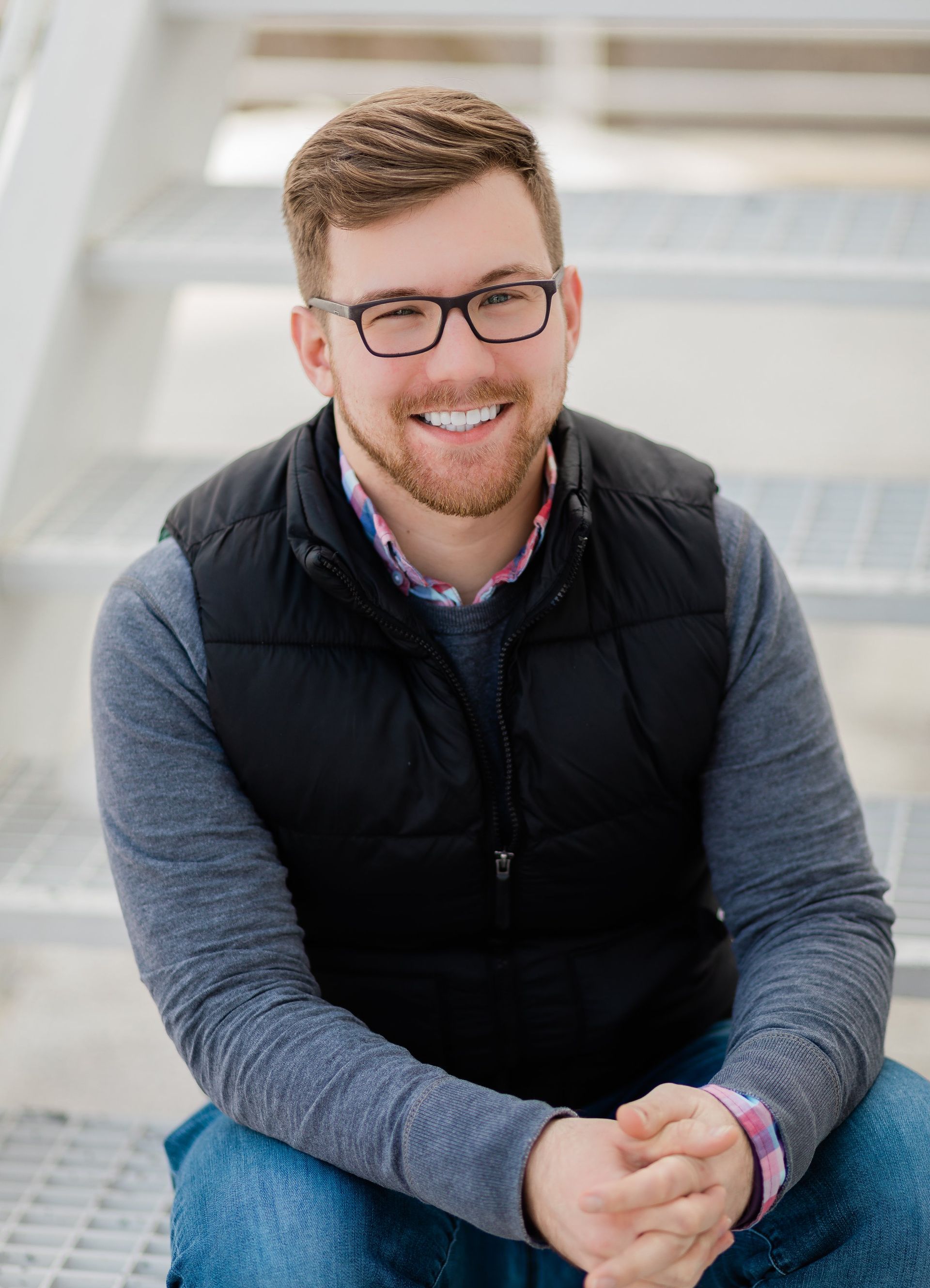 Man sitting on metal stairs, smiling. Wearing glasses, vest, and sweater.