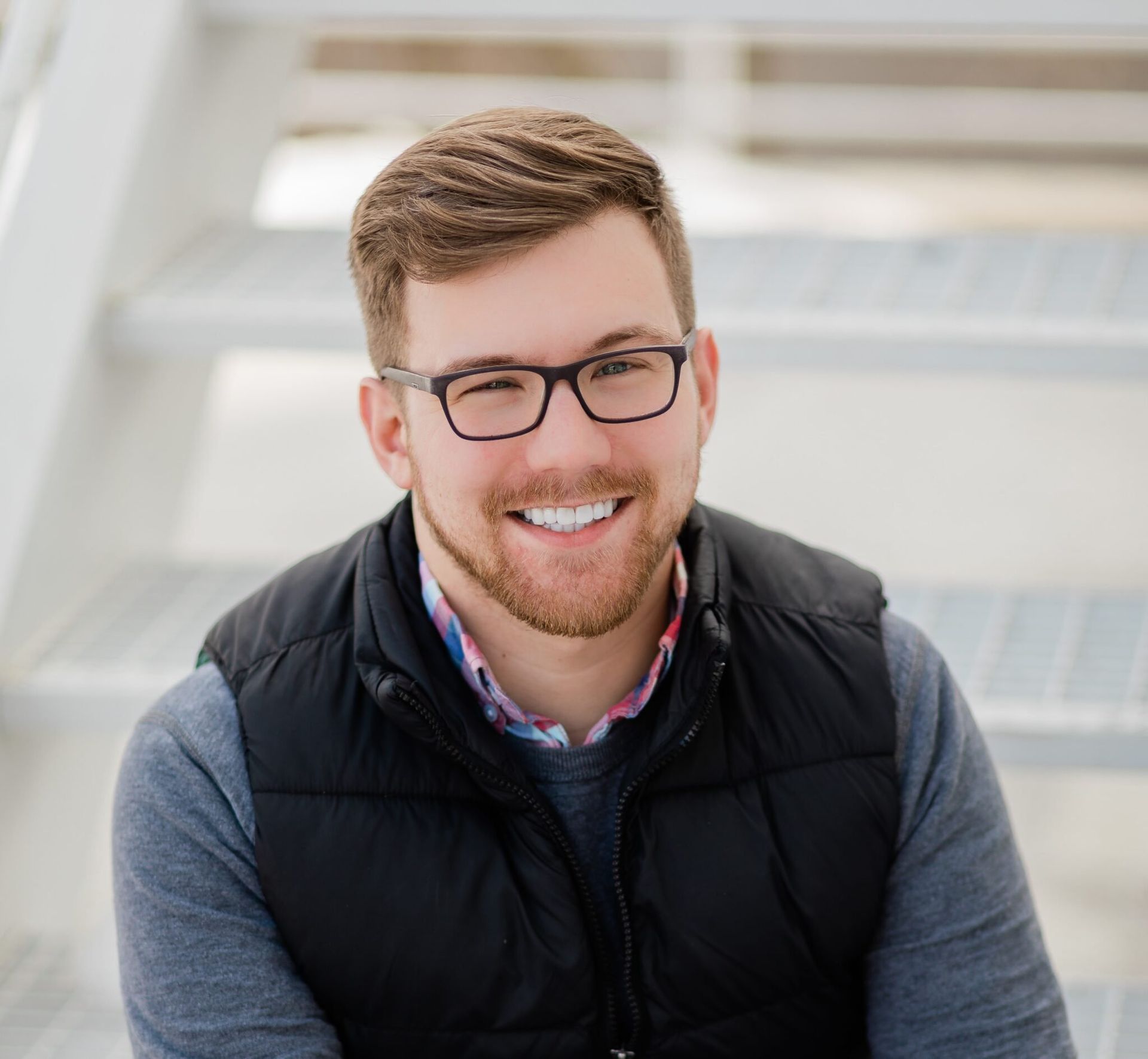 Man with glasses smiles, sitting on white stairs, wearing a black vest and gray sweater.