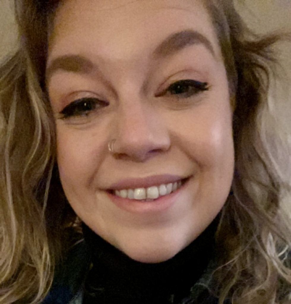 Smiling woman with curly blonde hair, wearing a nose ring and black turtleneck.