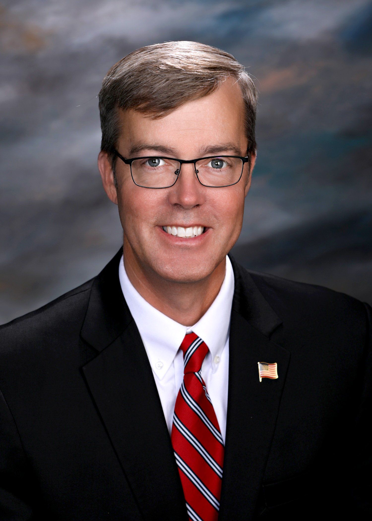 Man wearing glasses and a suit, smiling, with a red and white striped tie.
