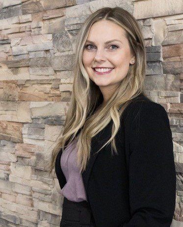 Woman in black blazer smiles, stands by a stone wall.