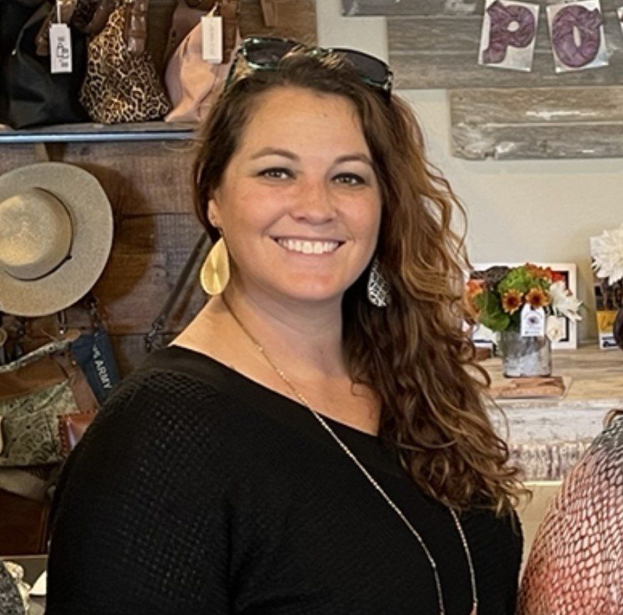 Woman with sunglasses on her head smiles, gold earrings, long wavy brown hair, black top, in a shop.