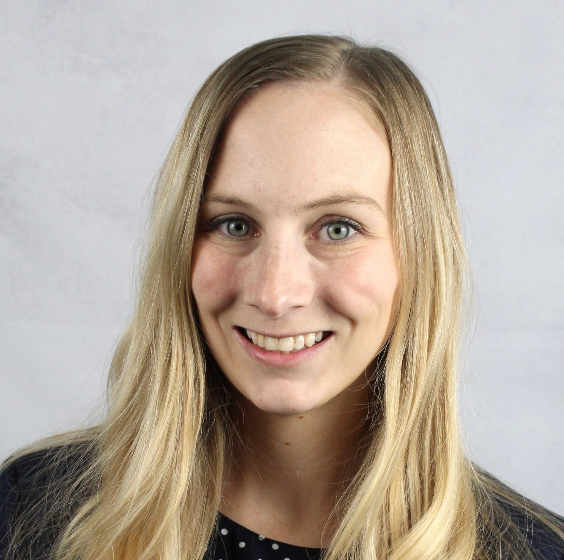 Woman with long blonde hair smiles at the camera, wearing a dark top with a polka dot pattern against a grey background.