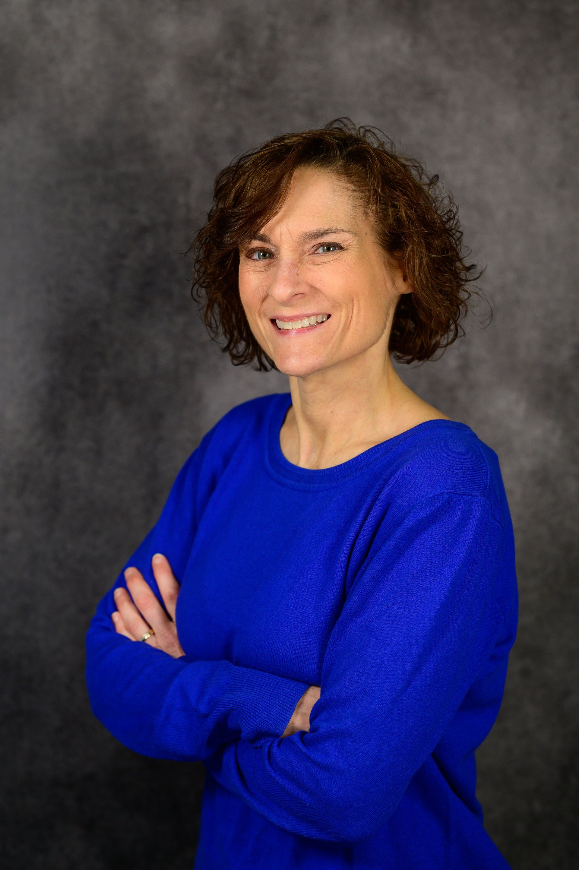 Woman with curly brown hair, arms crossed, wearing a blue sweater, smiling in front of a gray backdrop.