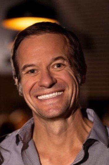 Man with a wide smile in a button-down shirt, lit from above, indoor setting.