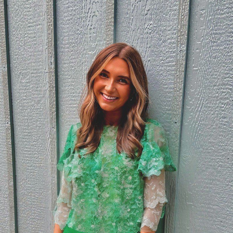 Woman with long, wavy blonde hair, wearing a green lace top, smiles in front of a blue wooden wall.