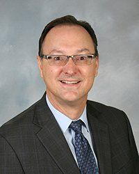 Man in a suit and tie, smiling, wearing glasses, in front of a gray gradient background.