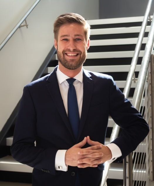 Man in a navy suit smiles at the camera, hands clasped. He stands near a staircase with black and white steps.