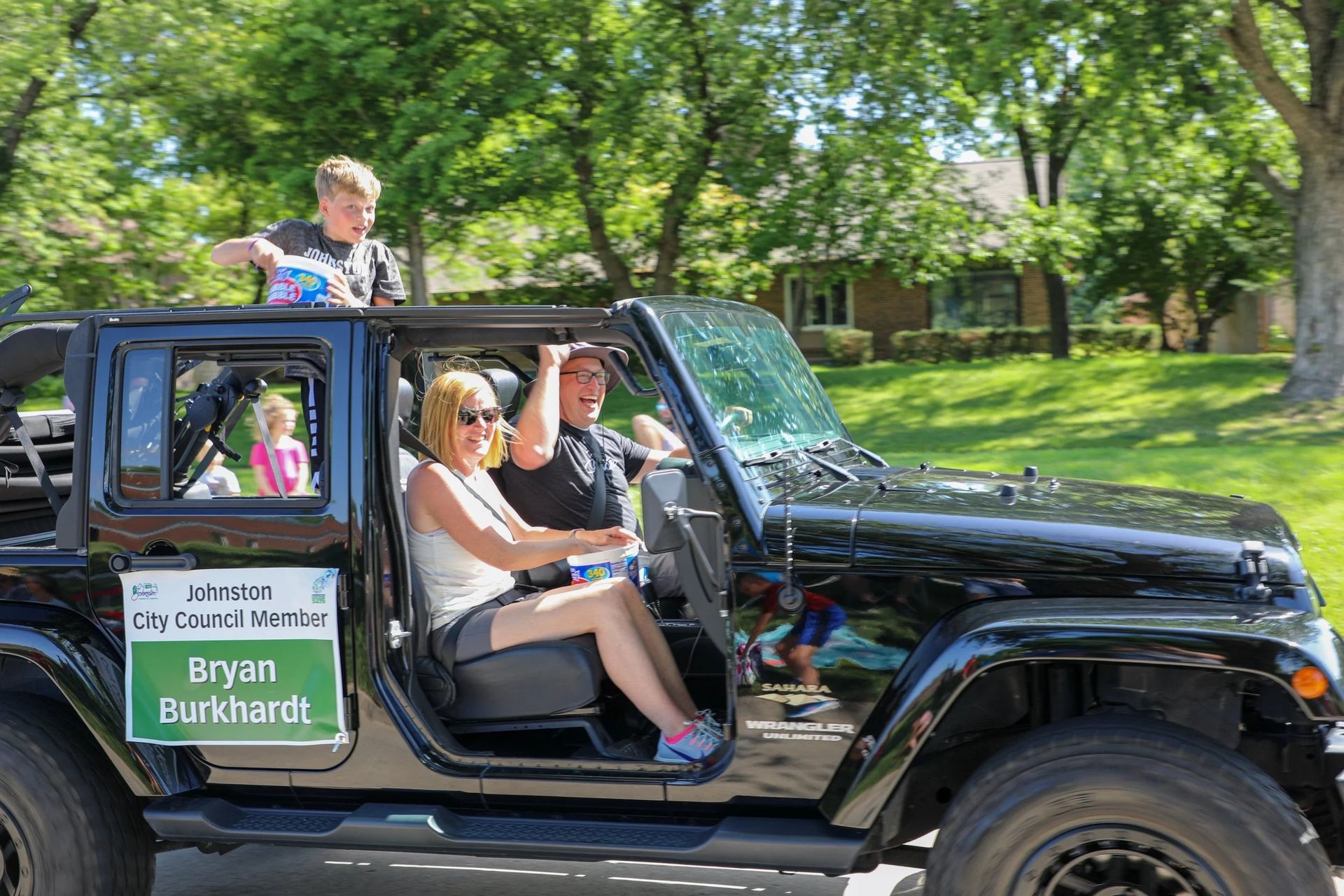 Family in a black Jeep in a parade: woman driving, man smiling, child on roof waving, child in backseat.