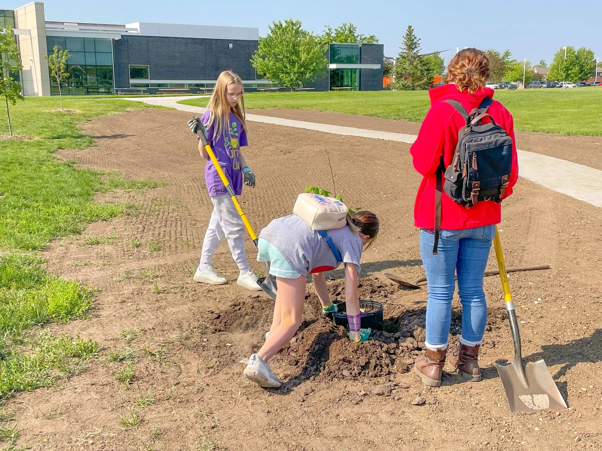 Three people planting in a garden bed. Two use shovels, one crouches planting. Exterior, sunny day.