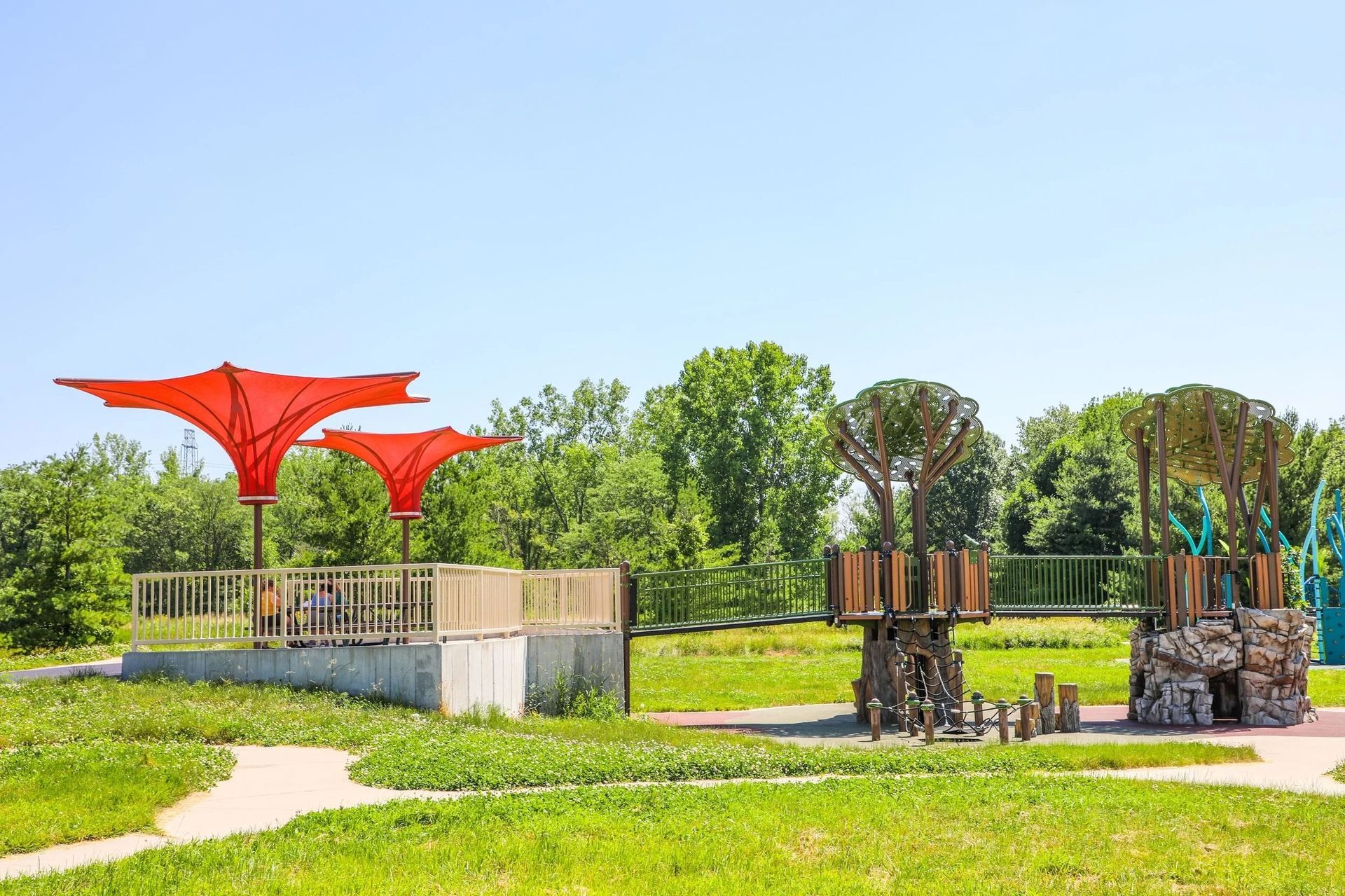 Playground with red shade structures, wood and stone climbing structures, and green grass.
