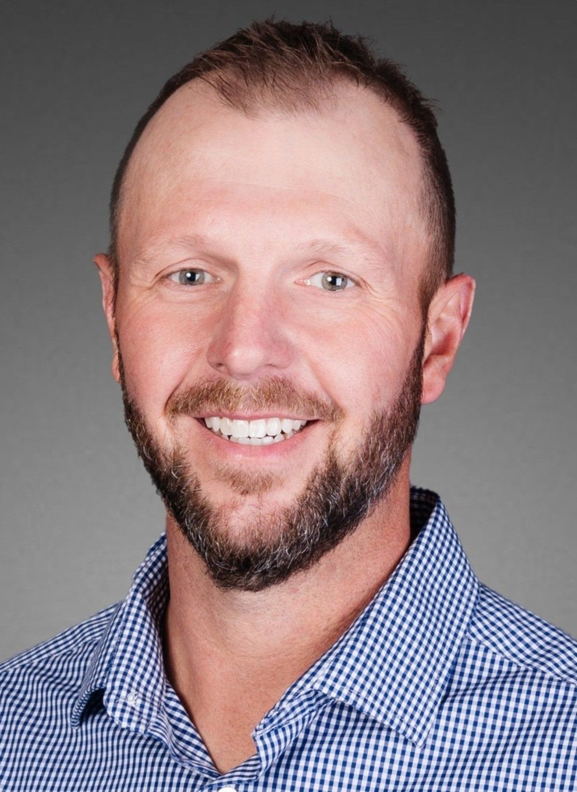 Man with a beard smiling, wearing a blue patterned button-down shirt, headshot against a gray gradient.
