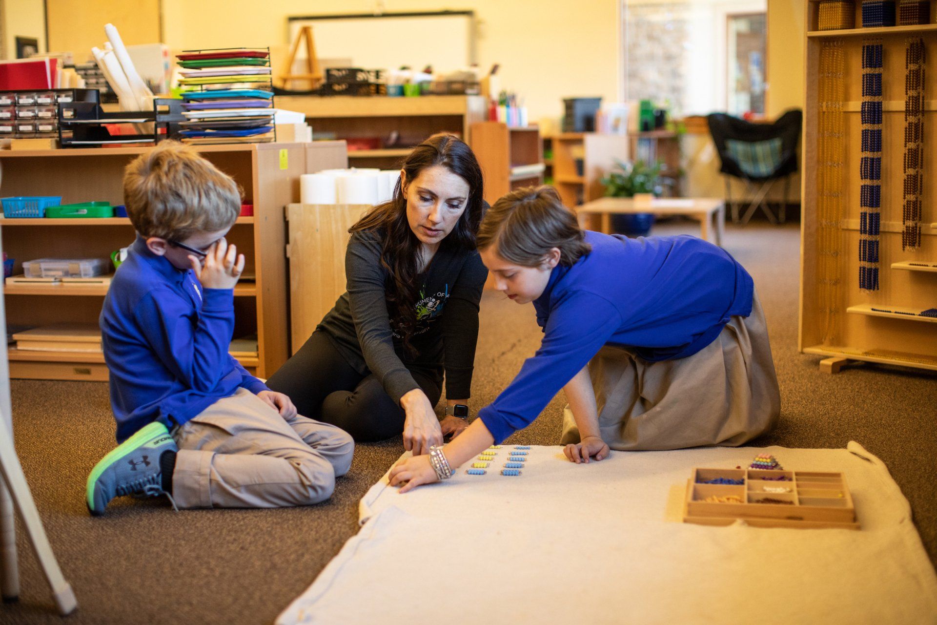 A montessori guide  is sitting on the floor with two children in a classroom.